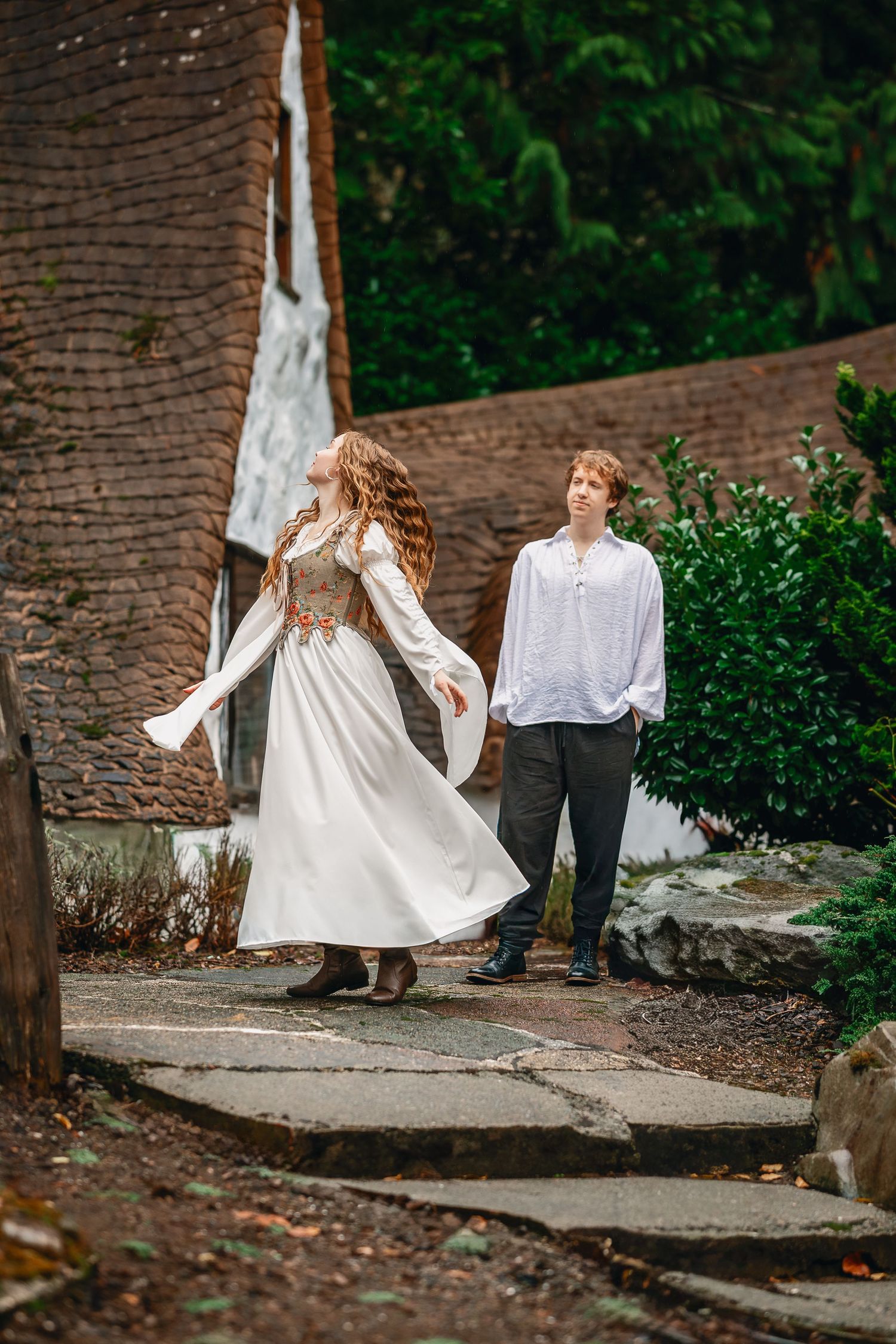 Two figures in white and dark clothing walk together along a stone path surrounded by greenery.