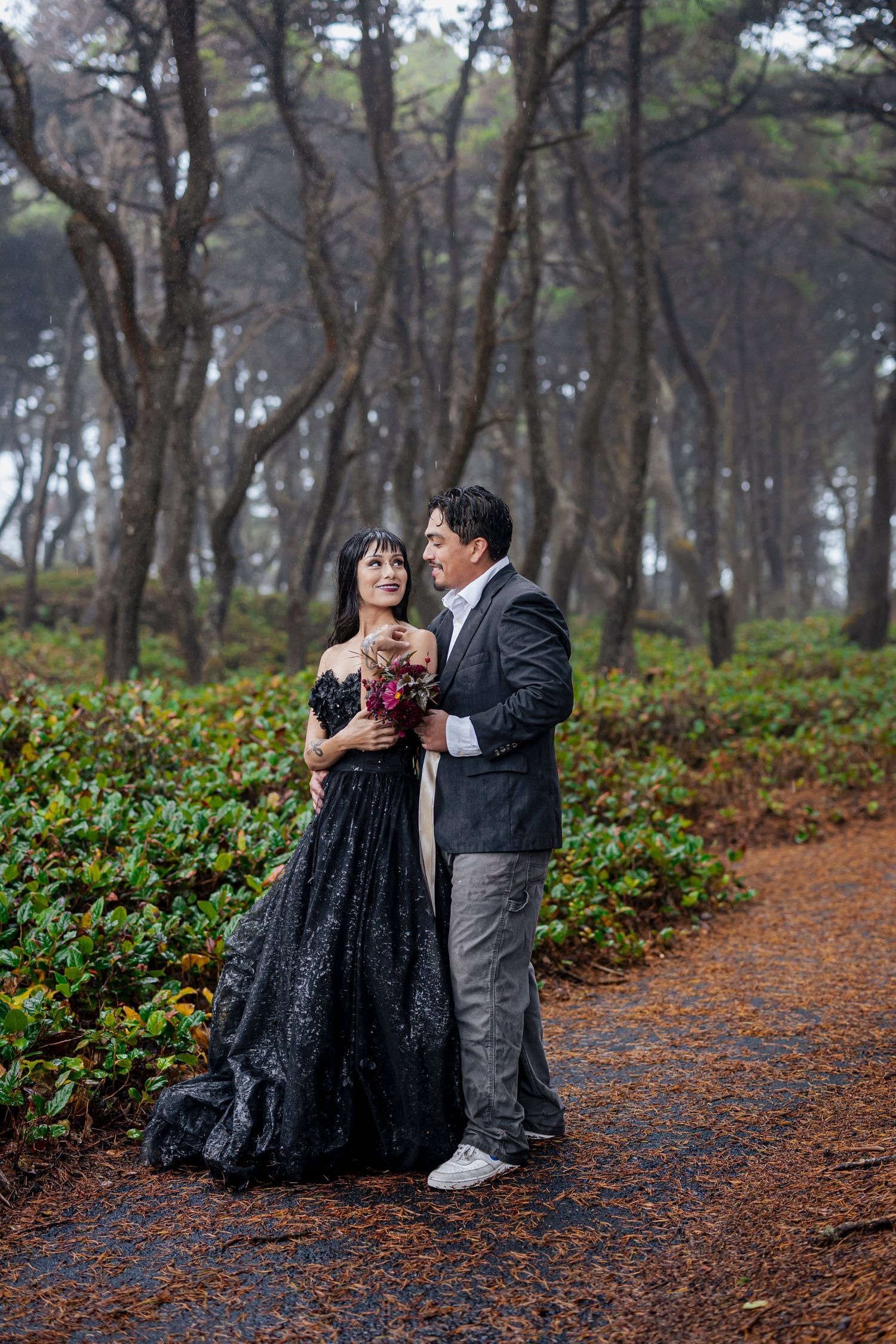 A couple in formal attire share a romantic moment on a misty forest path, with tall pine trees in the background.