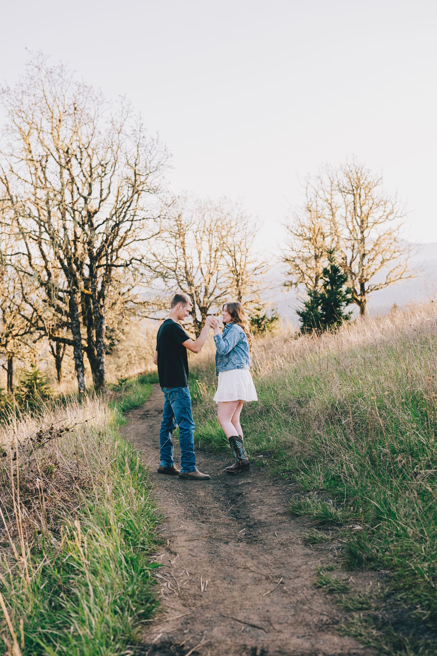 Two people standing together on a dirt hiking trail surrounded by barren trees during sunset.