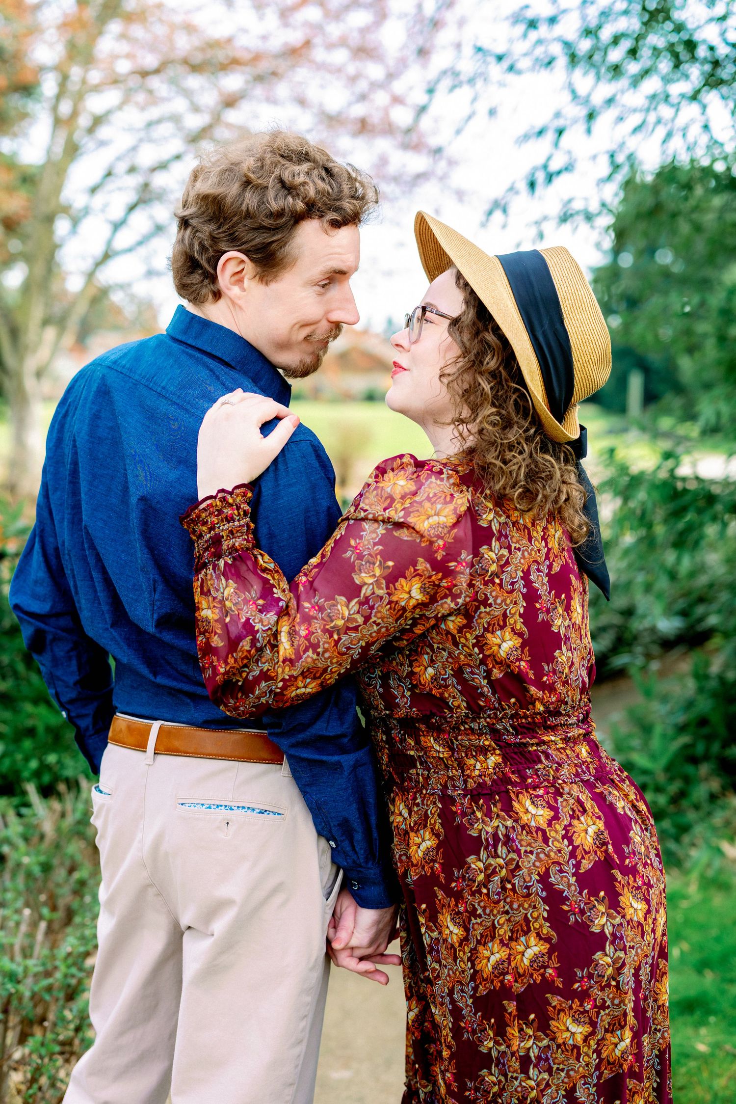 A romantic couple embraces outdoors, with one wearing a burgundy floral dress and the other in a navy shirt.