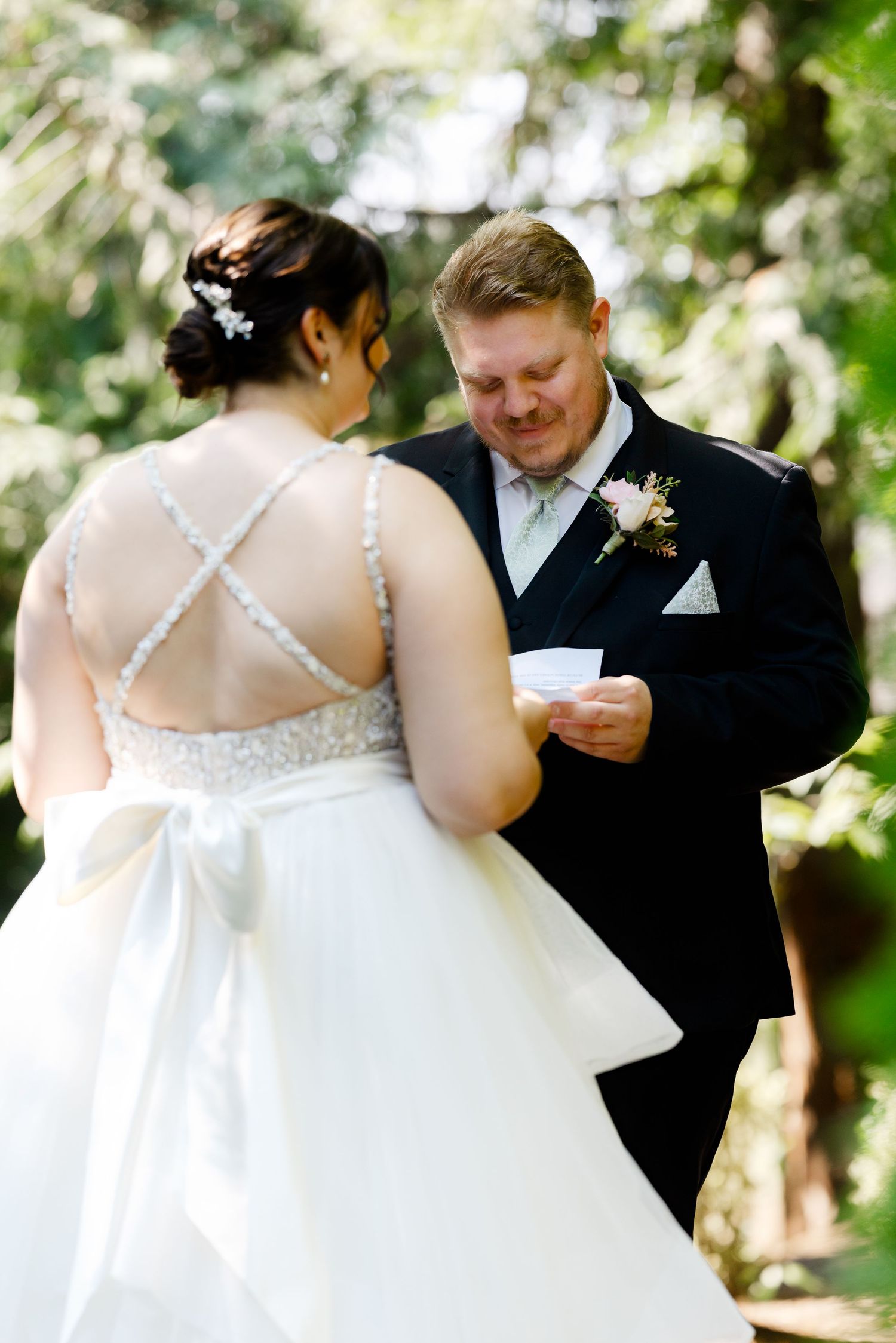 Groom reads vows from paper while bride in beaded backless gown listens outdoors.