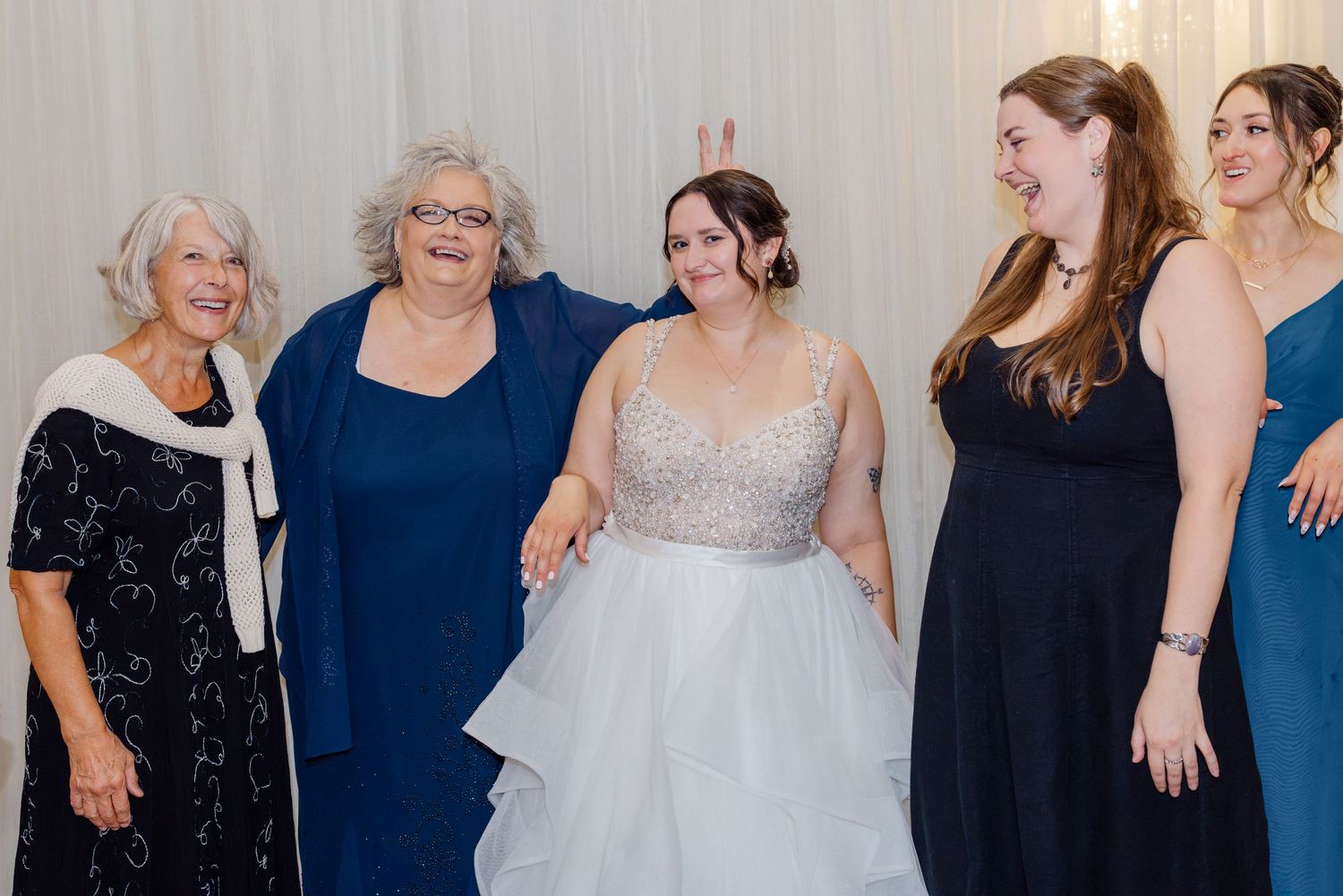 Bride smiling with four women in blue and black dresses at indoor wedding reception.