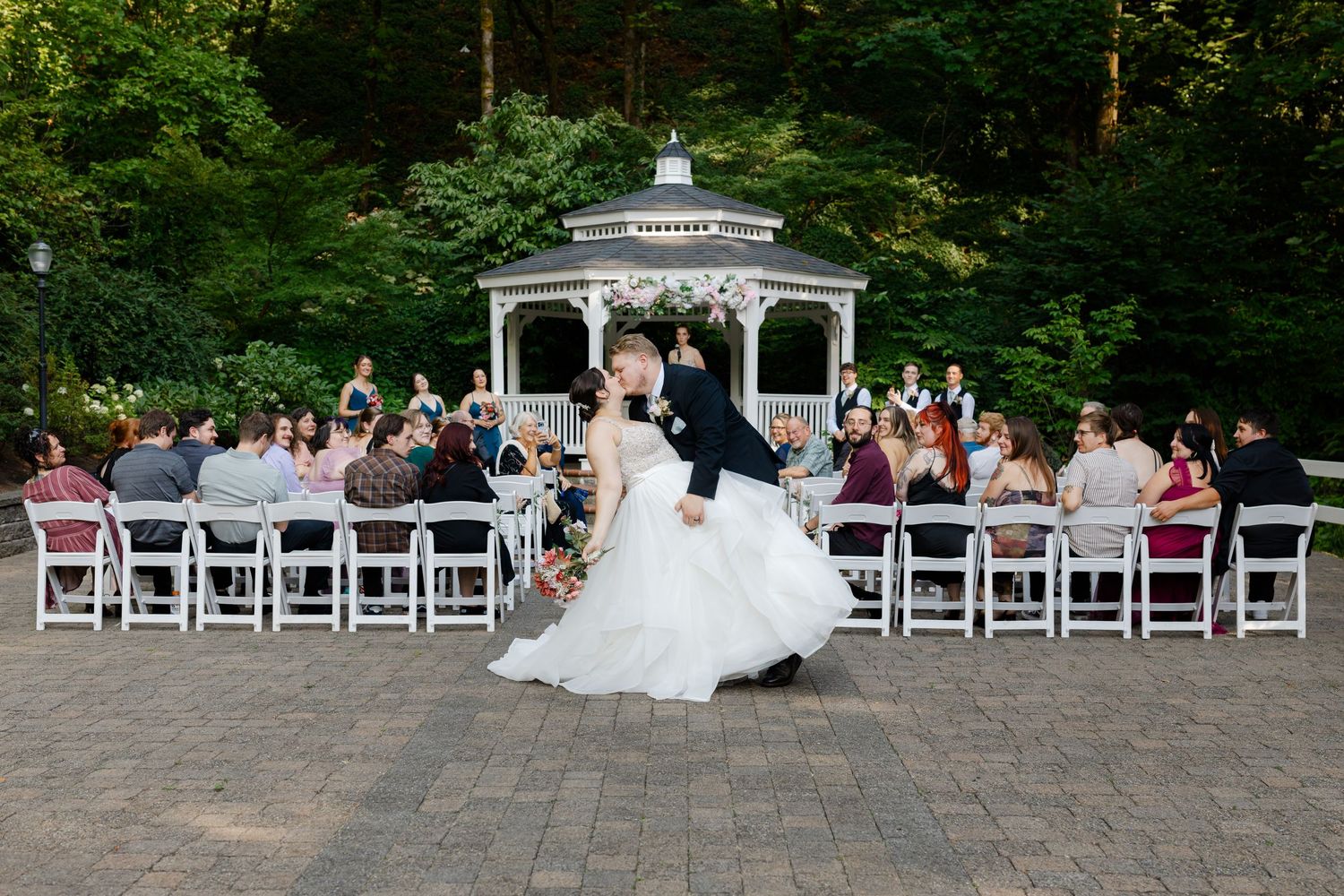 Bride and groom sharing a romantic dip kiss in front of seated wedding guests at outdoor gazebo.