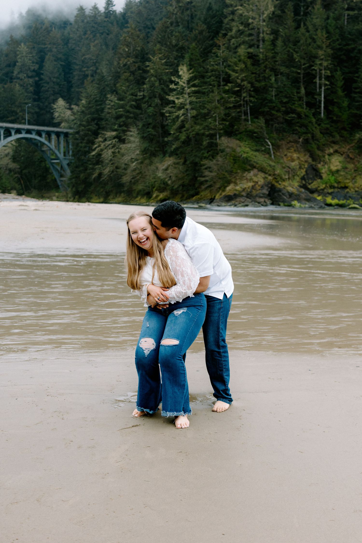 Couple laughing and embracing barefoot in ocean waves on Oregon beach near historic bridge.