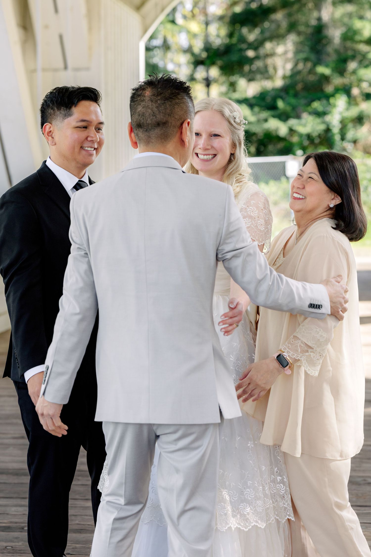 A joyful wedding party greets each other with hugs and laughter on a covered wooden bridge outdoor venue.