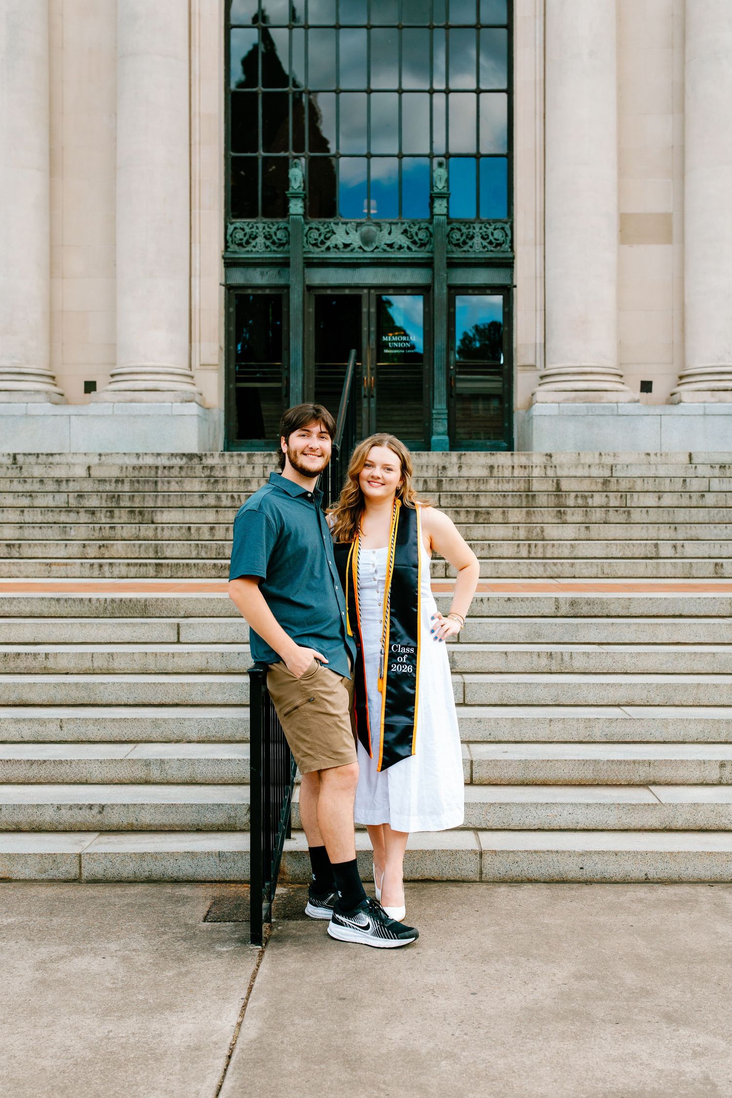 Female graduate in white dress with gold stole poses with male companion on university building steps.