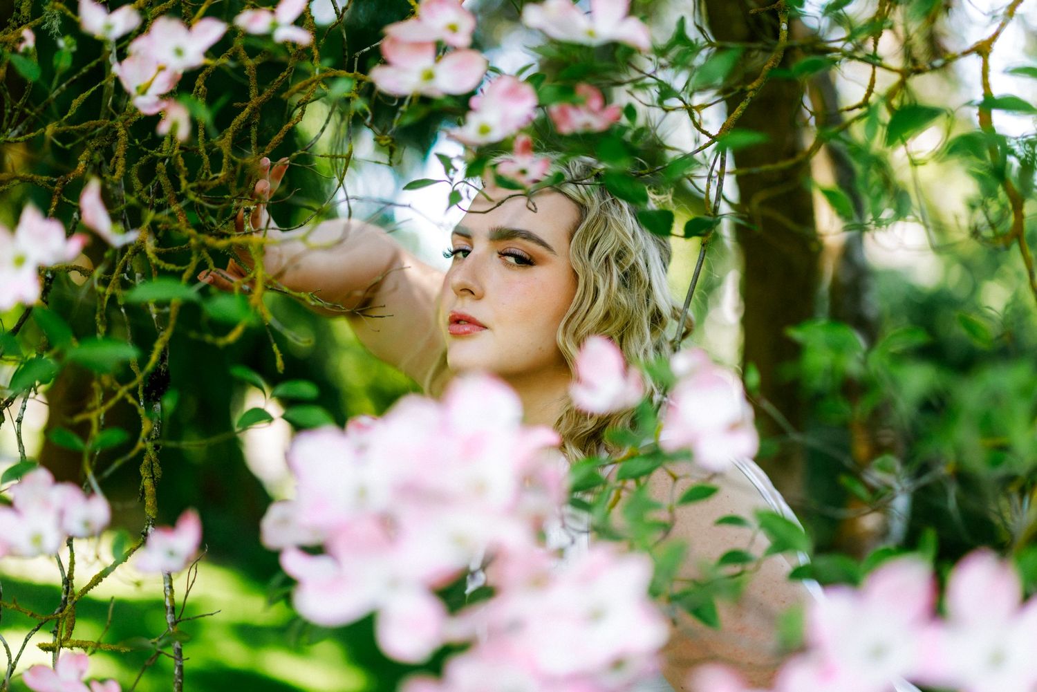 A young blonde woman poses among blooming pink dogwood flowers in a lush green garden setting.