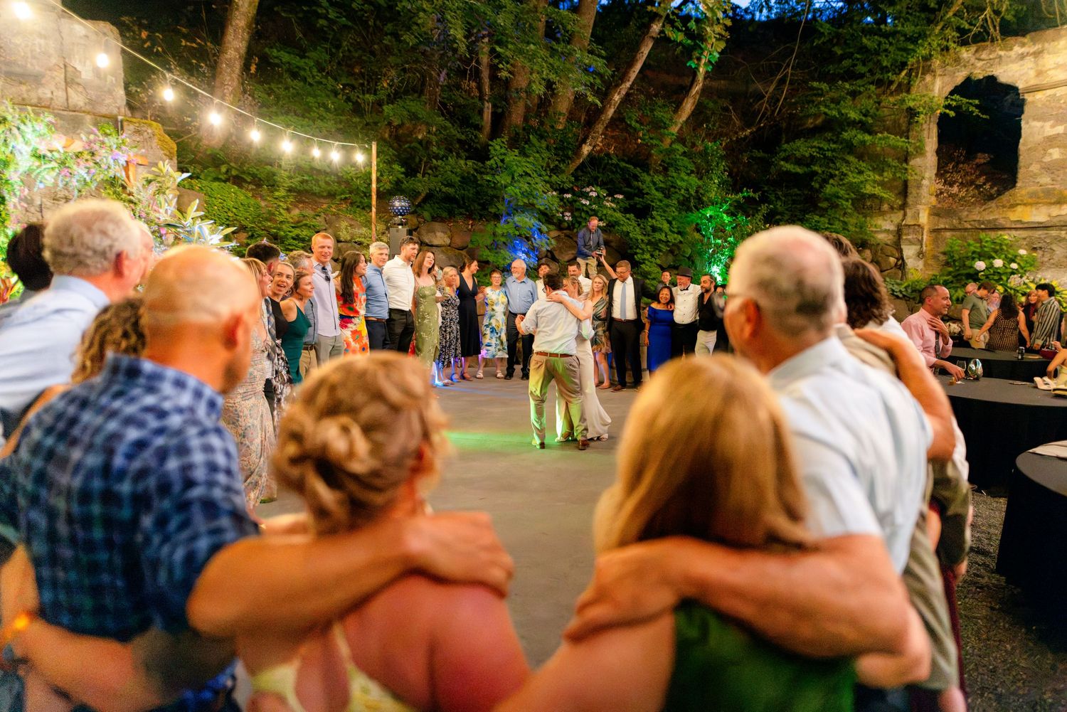 Wedding guests gather on dance floor under string lights during evening outdoor reception surrounded by lush greenery.
