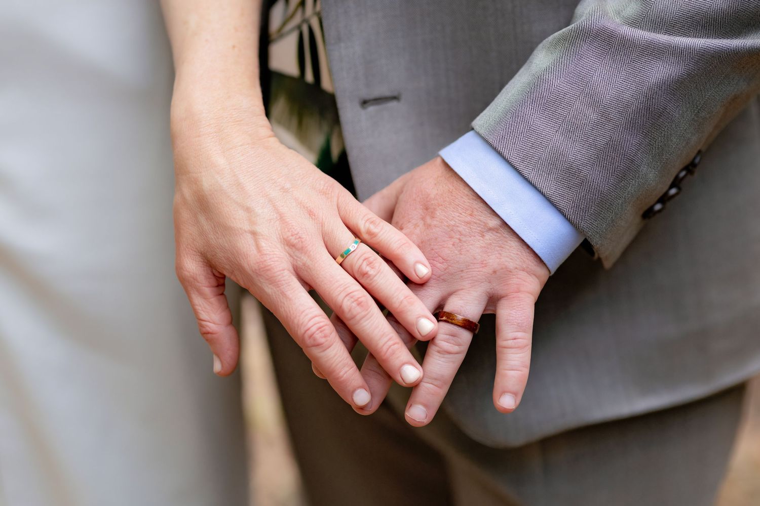 Close-up of newlyweds holding hands showing wedding rings, bride in white dress and groom in gray suit.