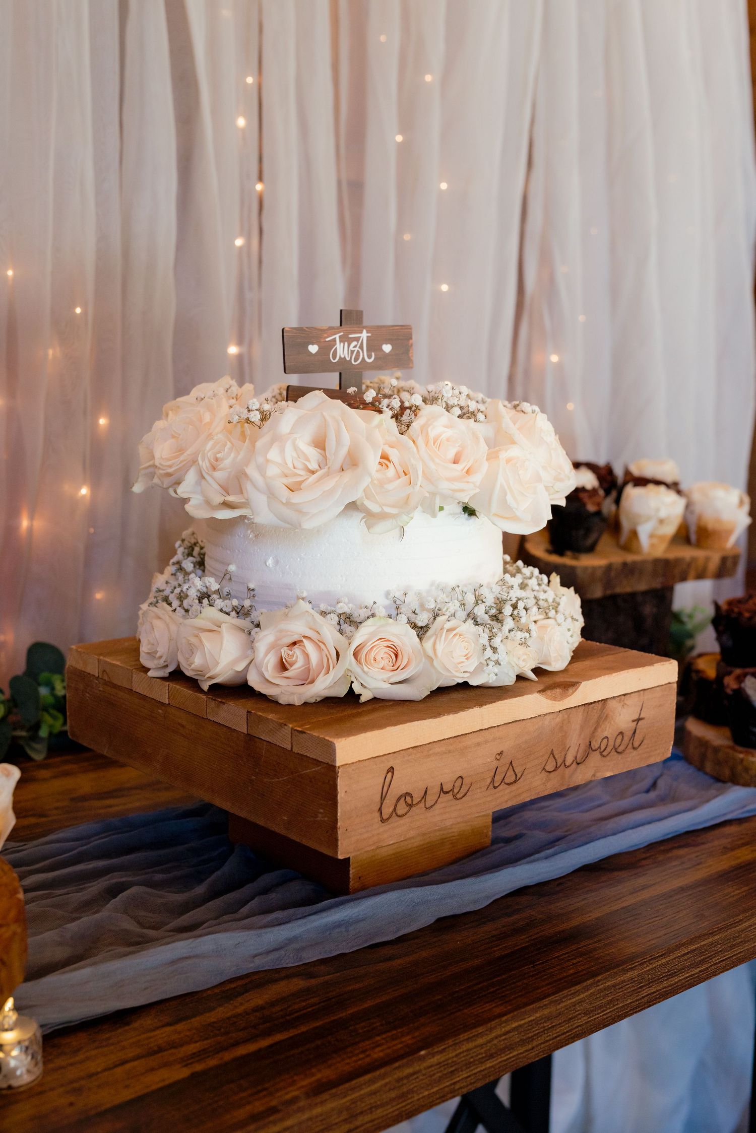 White wedding cake adorned with blush roses on a wooden stand engraved with love is sweet, set against a twinkling light backdrop.