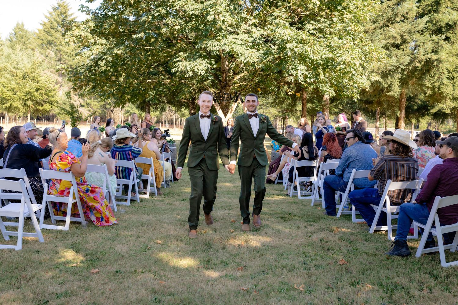 Two grooms in matching dark green suits hold hands while walking back down the aisle after their ceremony.