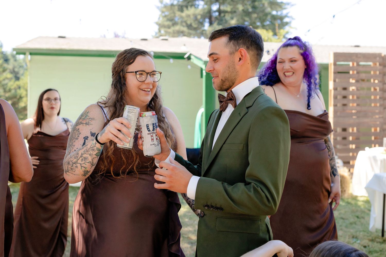 Bride and groom toasting with canned drinks at outdoor wedding, bridesmaids in brown dresses in background.