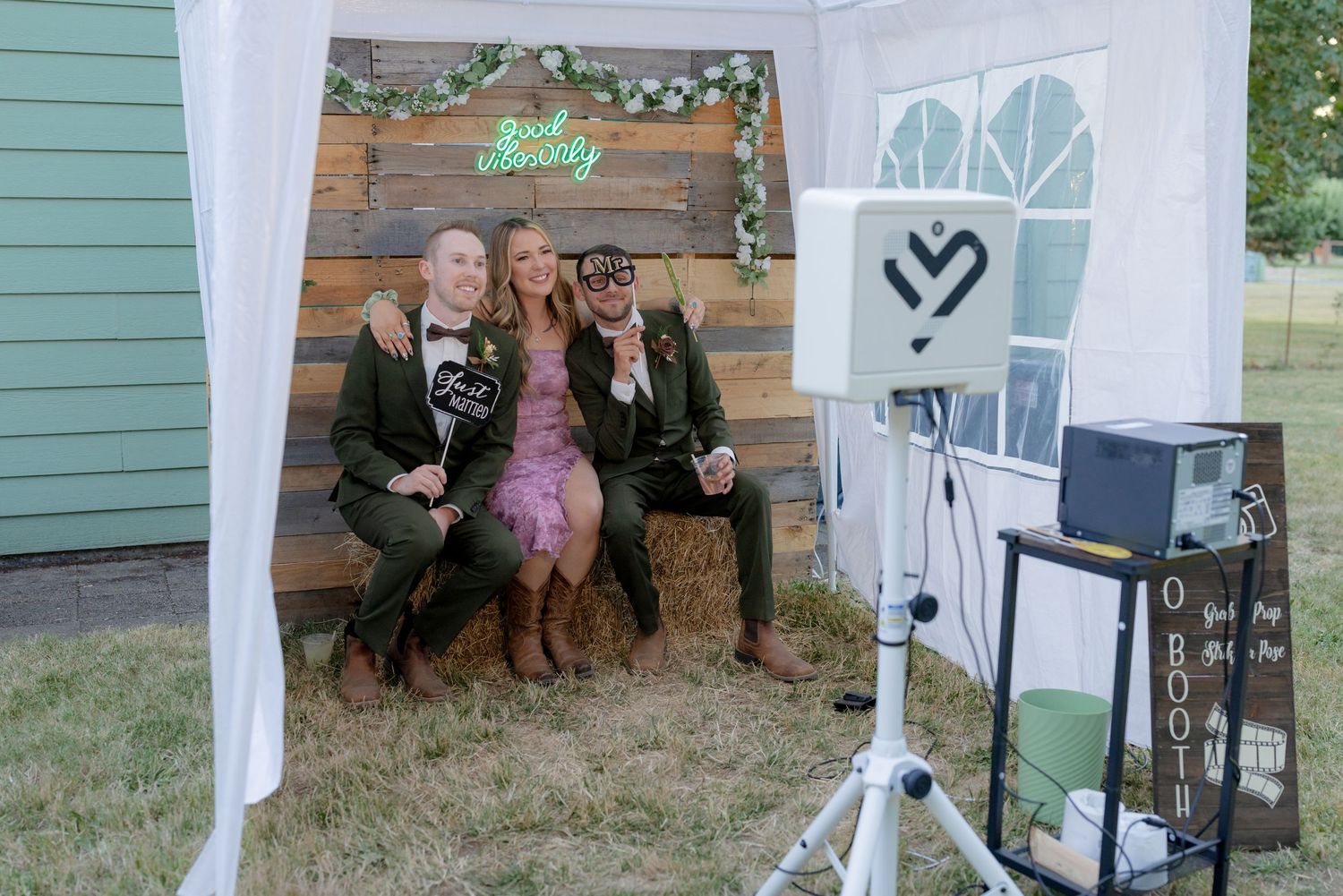 Three guests pose and smile at a rustic outdoor wedding photo booth with neon sign.