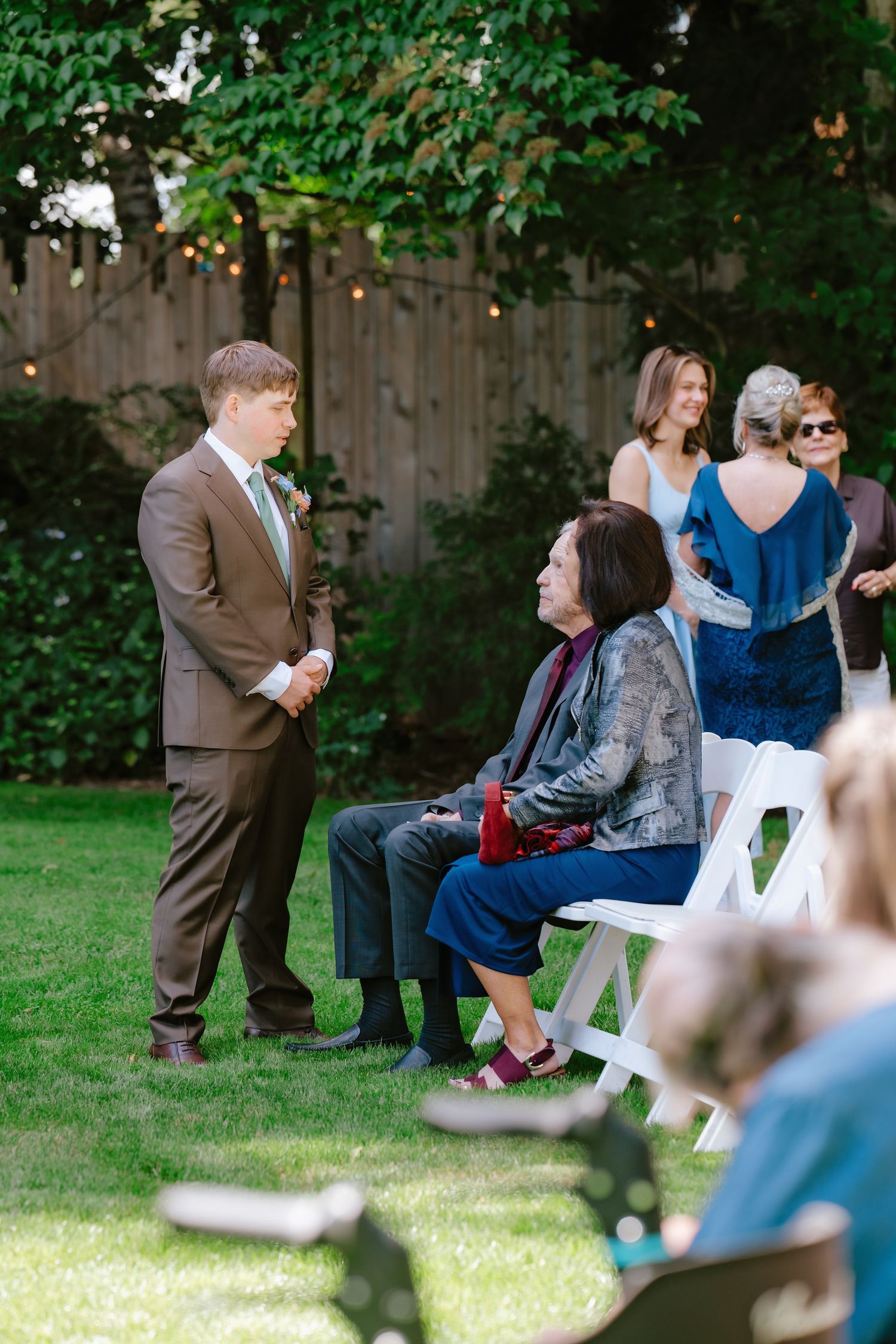 Groom in brown suit greeting seated guests at an outdoor backyard wedding ceremony on a sunny day.