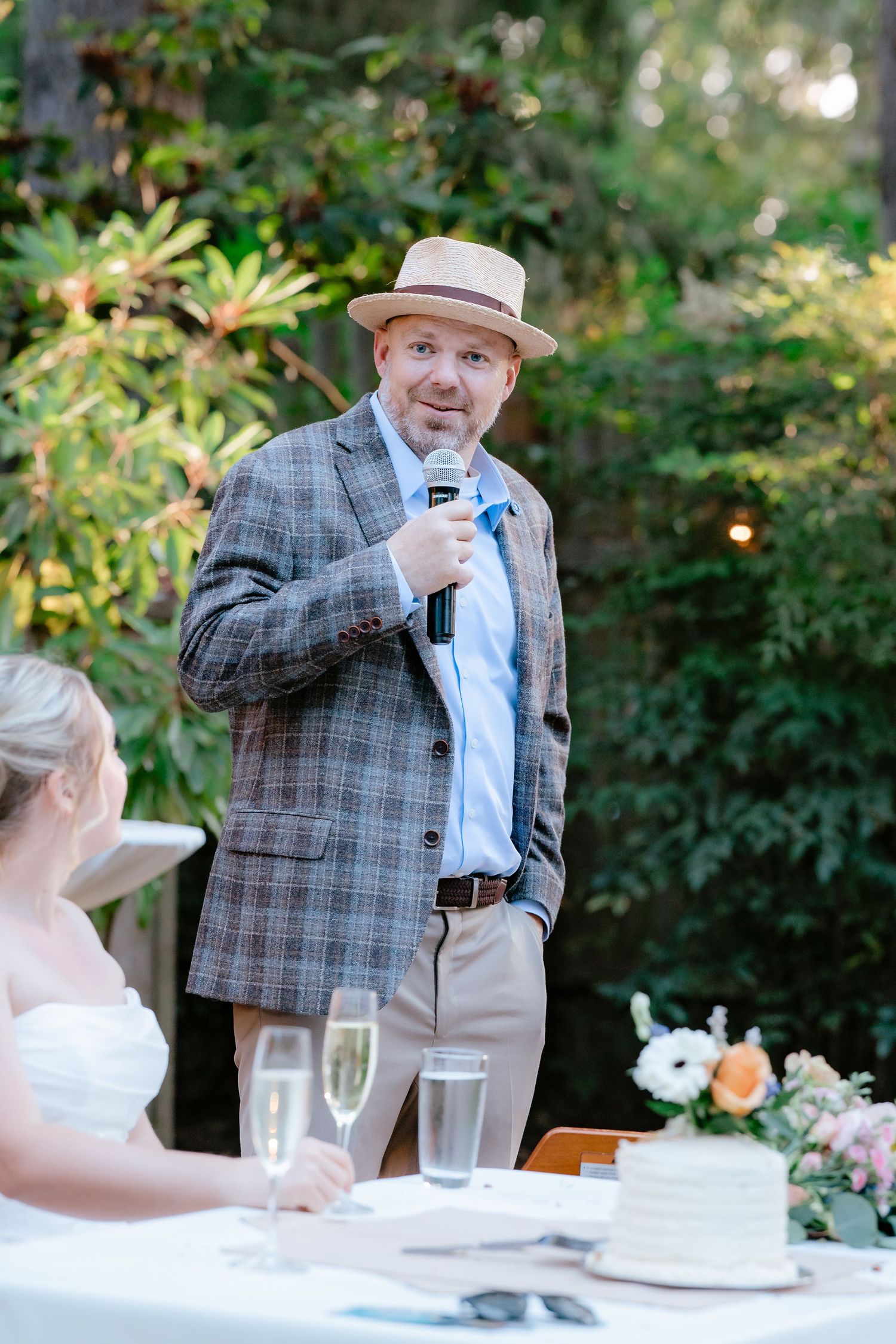 Man in plaid blazer and straw hat giving a speech at an outdoor wedding reception with microphone.