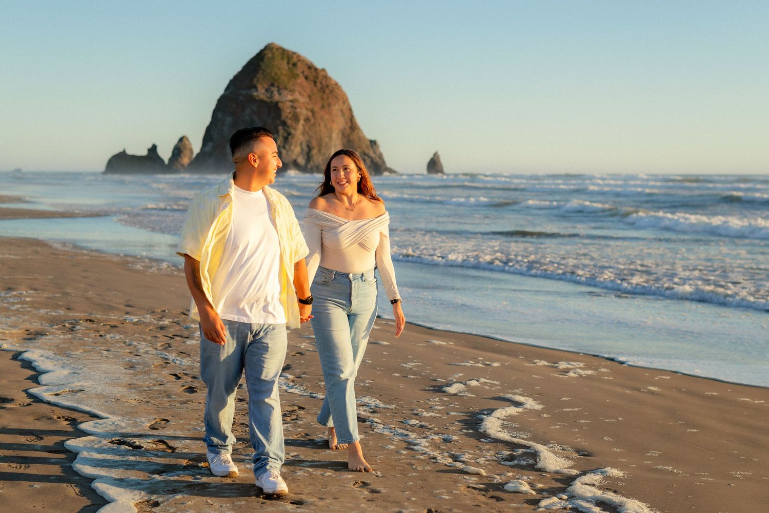 Couple walking hand in hand on a sandy beach at sunset with Haystack Rock in the background at Cannon Beach, Oregon.