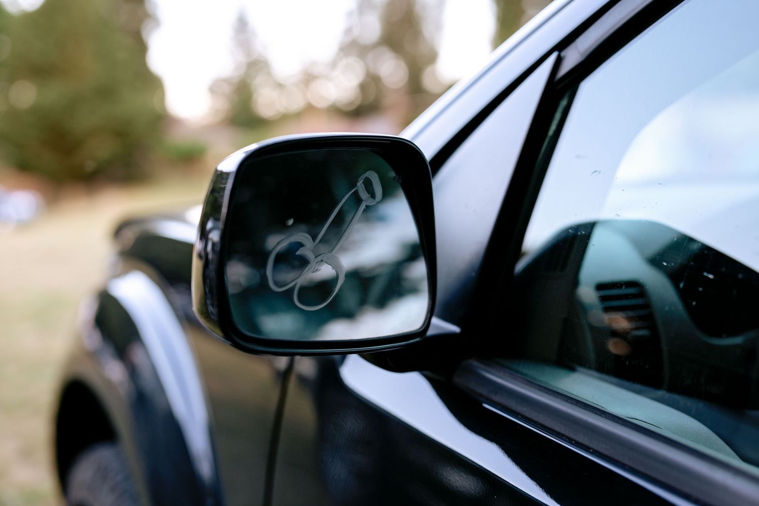 Close-up of a black car side mirror reflecting a decorative key ornament in a lush outdoor setting.