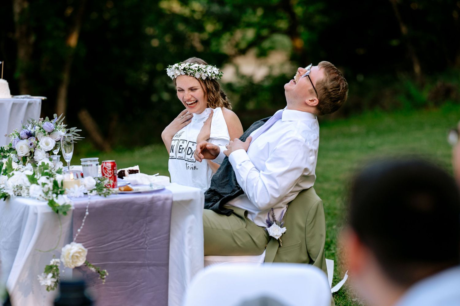 Bride in white dress with floral crown laughs with groom at decorated wedding reception table outdoors.