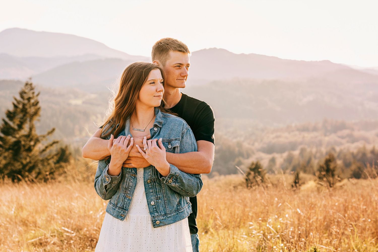 Couple embracing in golden field with mountain backdrop during sunset, woman in denim jacket, man in black shirt.