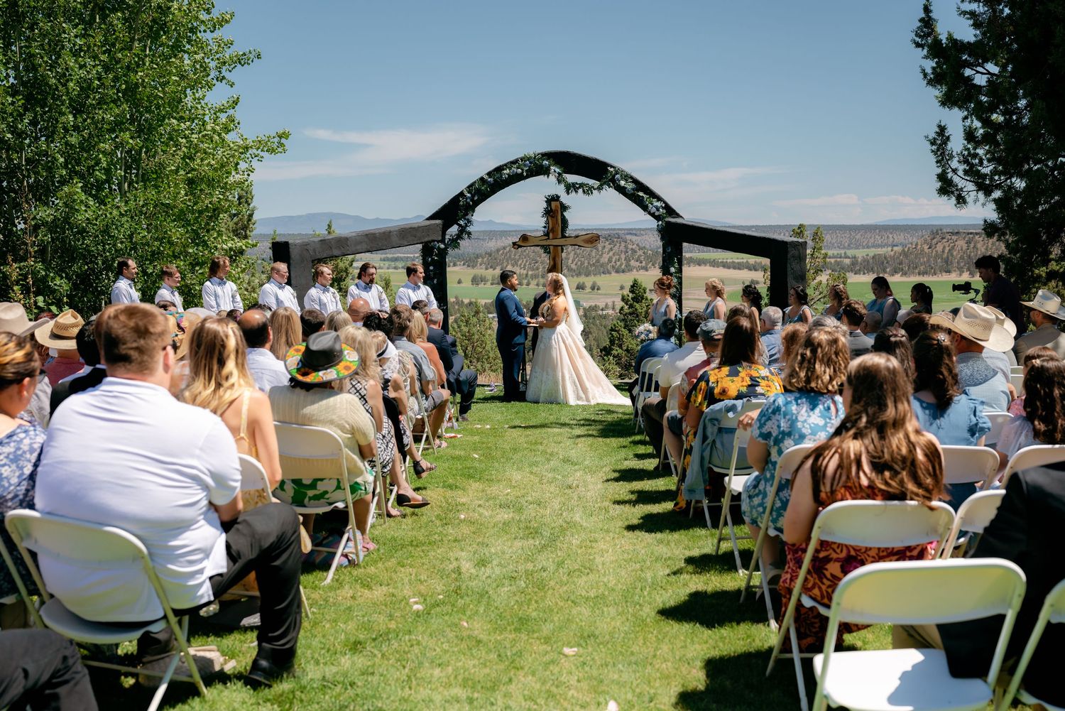 Guests seated in white chairs watch bride and groom exchange vows under rustic arch outdoors.