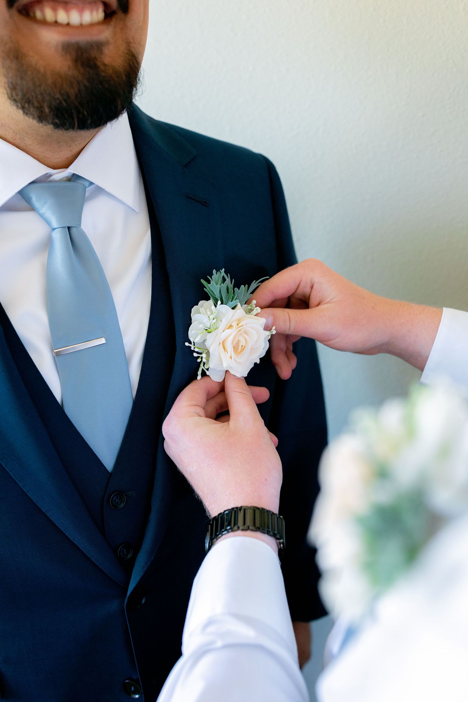 Close-up of hands pinning a white rose boutonniere onto a groom's navy suit lapel before the wedding.