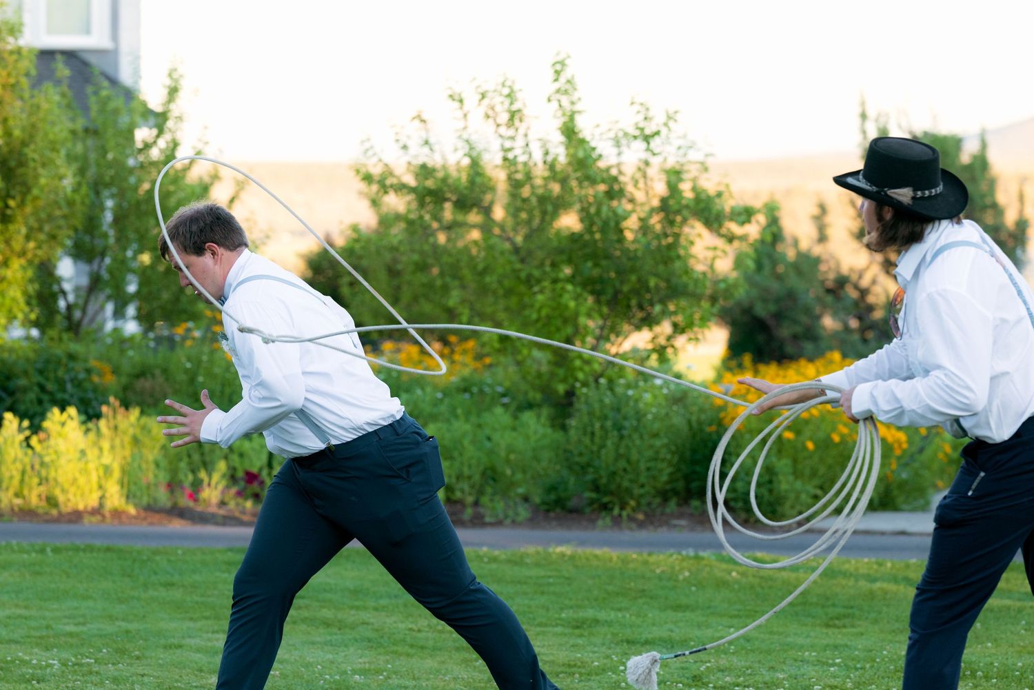 Two wedding guests in white shirts playfully lasso roping on a green lawn during a wedding reception.