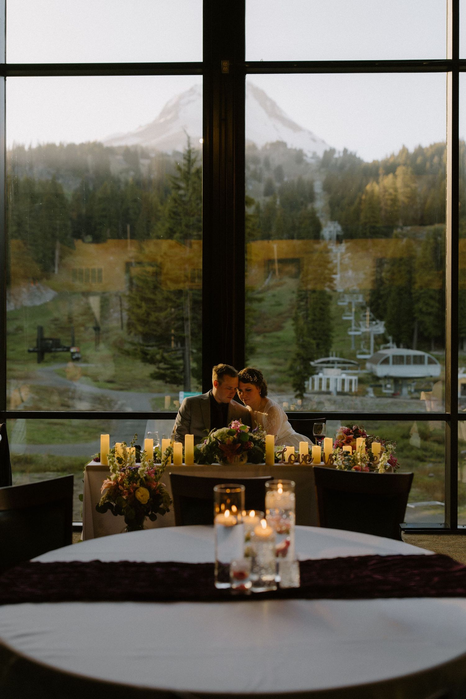 Couple sharing an intimate moment at sweetheart table with candles and mountain view through large window.