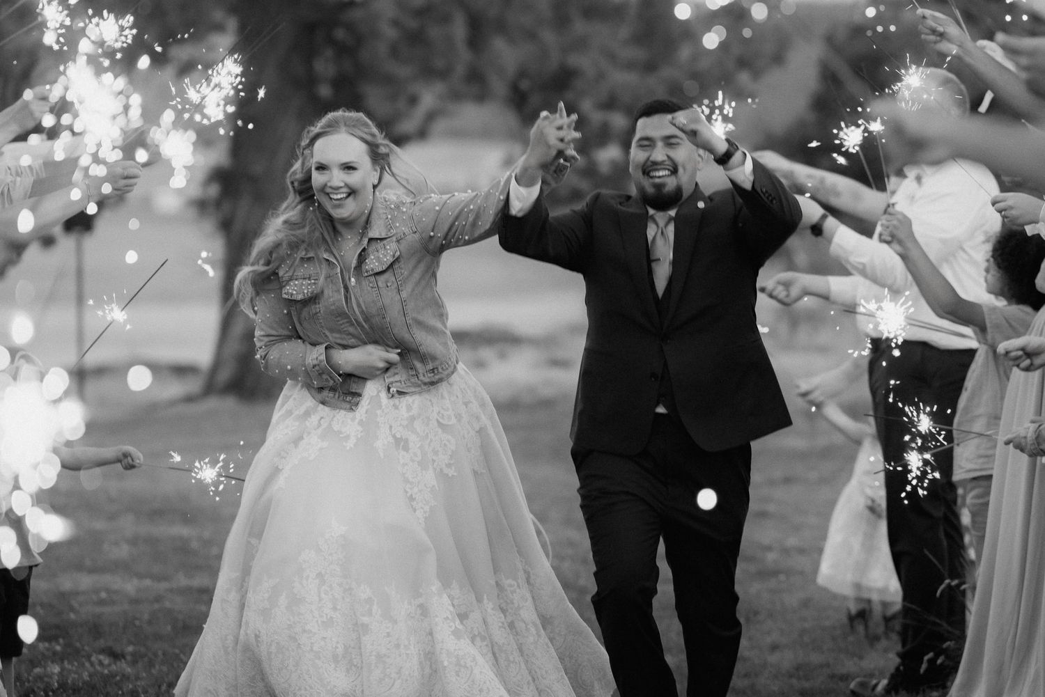 Black and white photo of a joyful bride and groom walking through a sparkler send-off at their outdoor wedding.