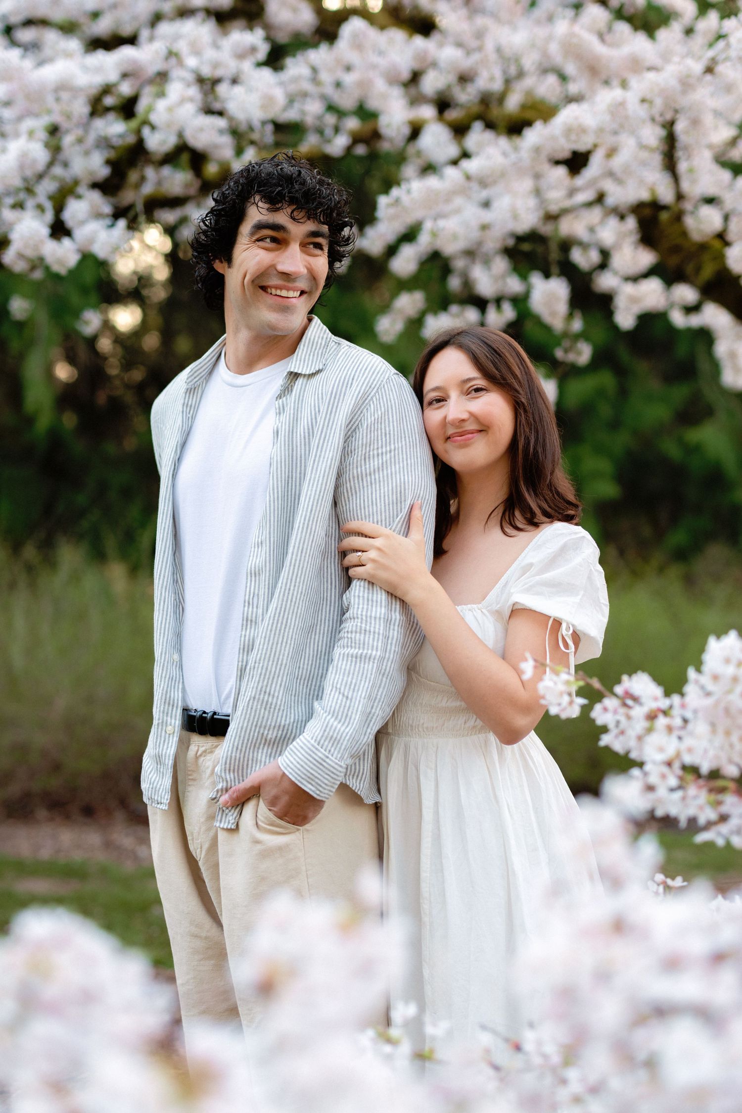 A couple poses together among blooming cherry blossom trees in springtime wearing coordinated white outfits.