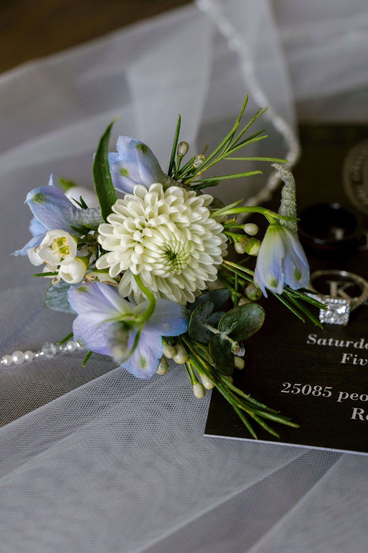 Delicate floral boutonniere with white dahlia and blue delphinium flowers on sheer fabric.