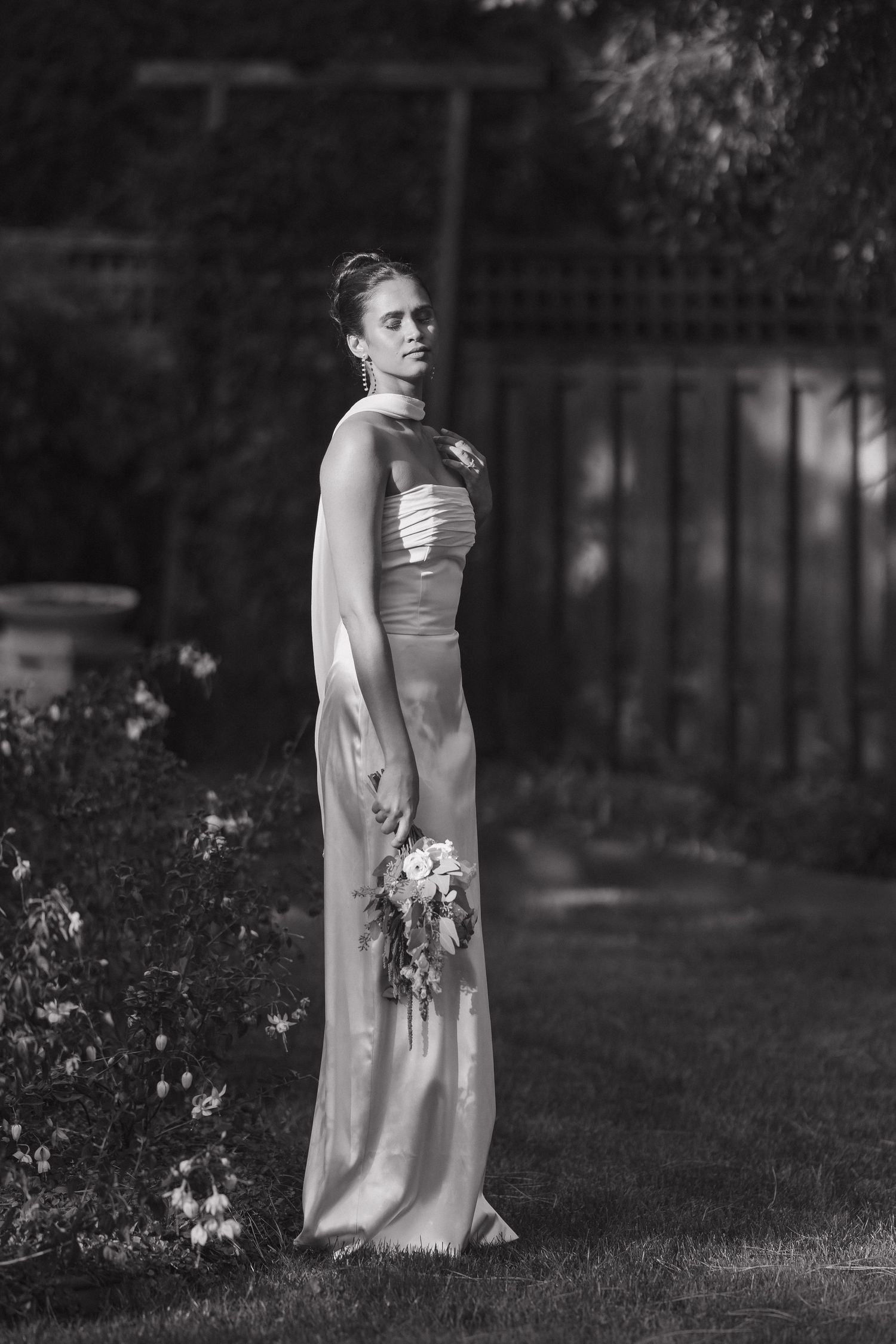 Black and white portrait of a bridesmaid in a formal gown holding a small bouquet in a garden setting.