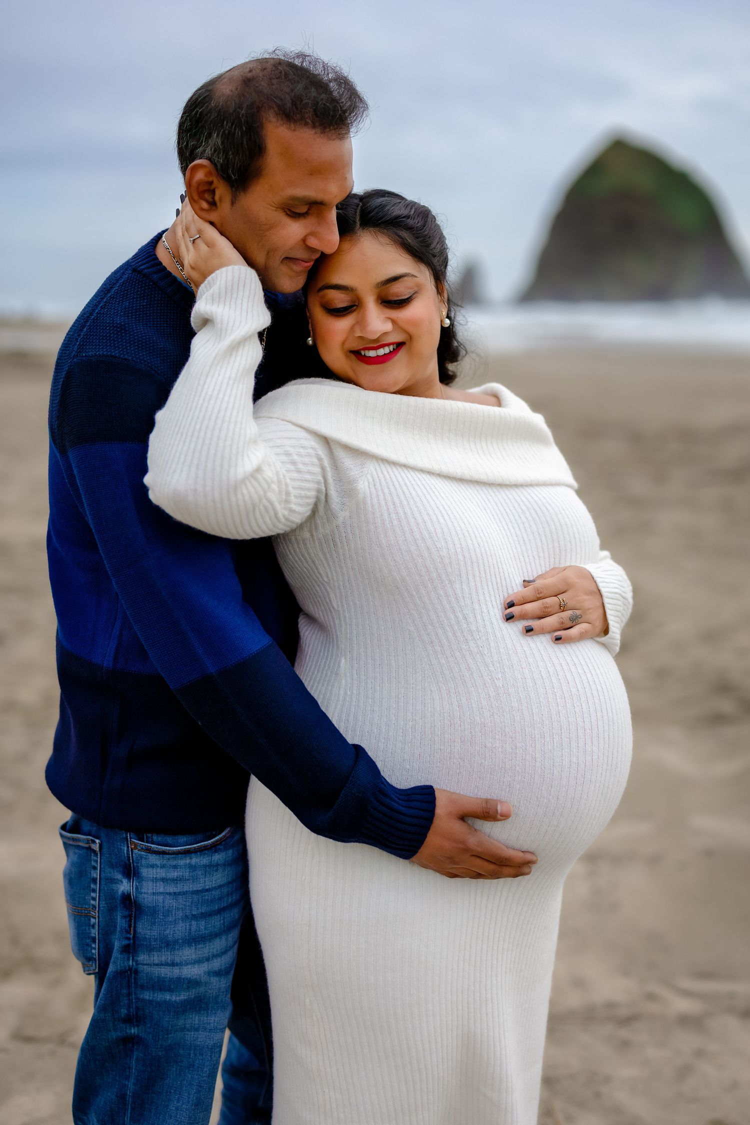 A couple embraces on the beach, with one person wearing a white sweater and the other in navy blue and jeans.