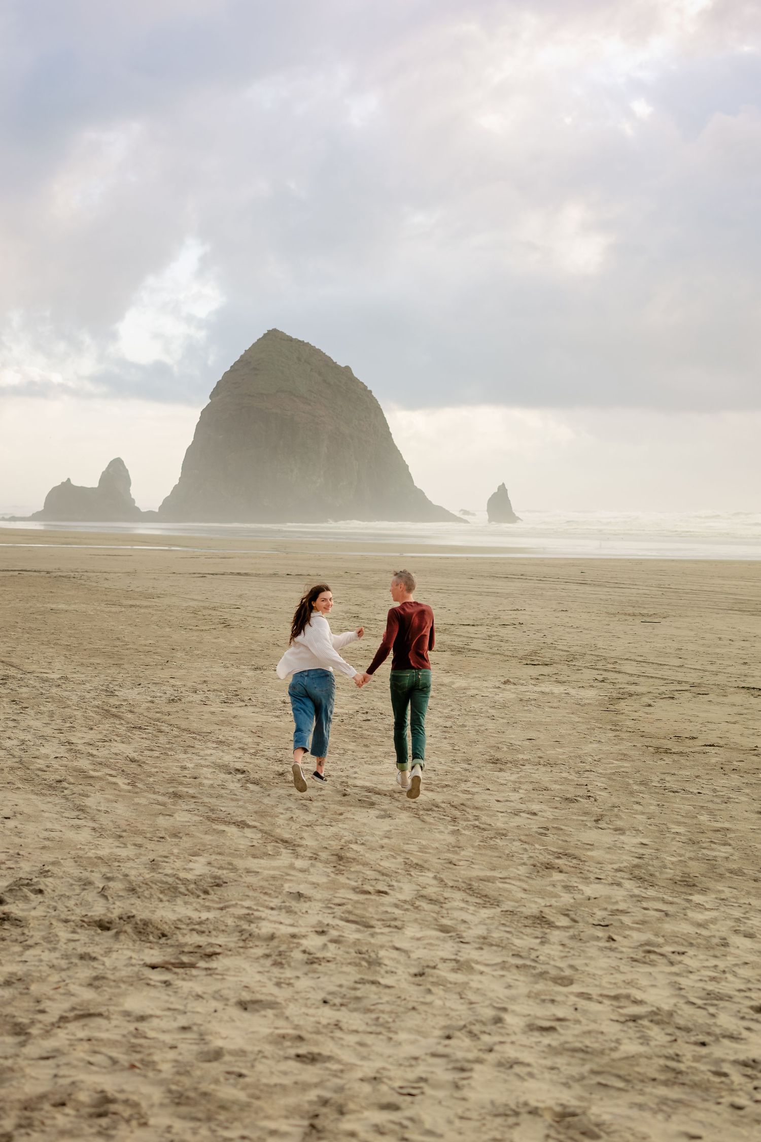 Two people walk together on a misty beach with dramatic rock formations visible in the distance at Cannon Beach, Oregon.