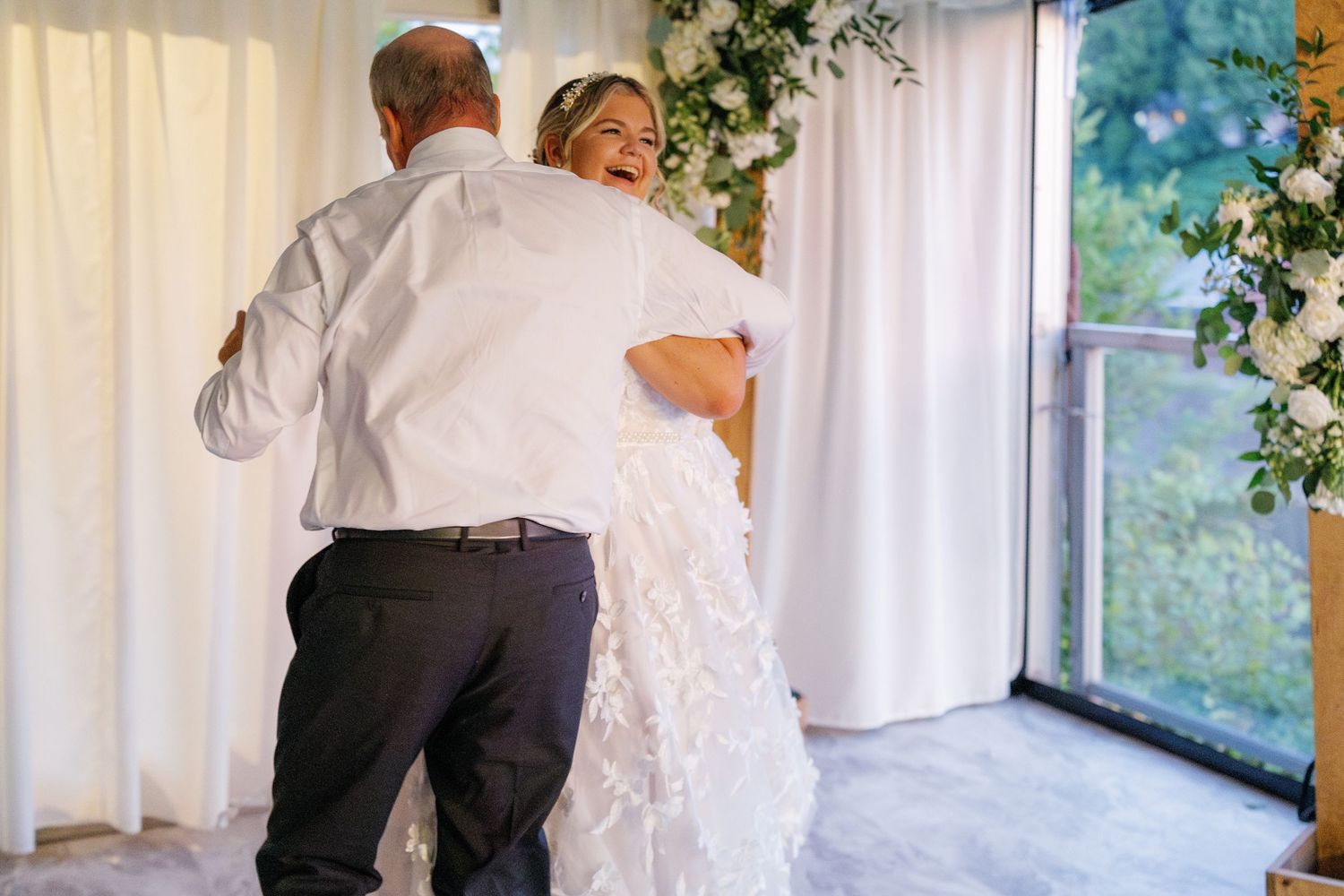 Wedding guests dance together in a bright sunlit reception hall with white curtains and greenery decorations.