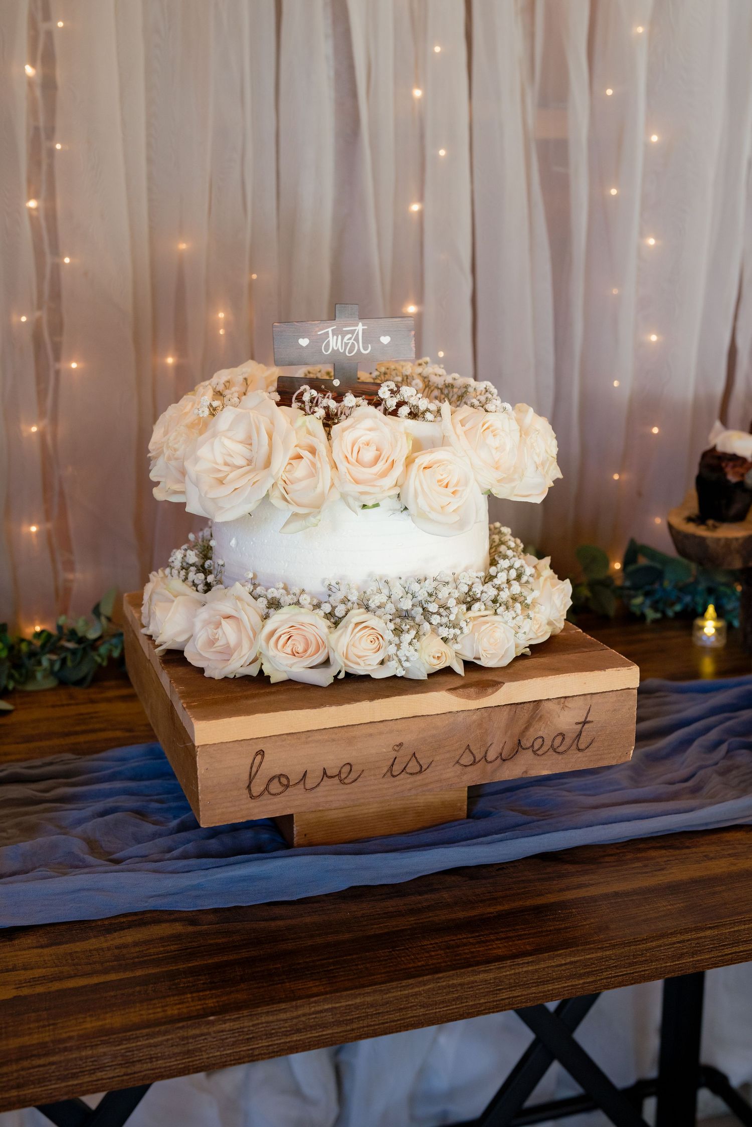 Elegant two-tier white wedding cake adorned with cream roses on a wooden stand reading love is sweet.