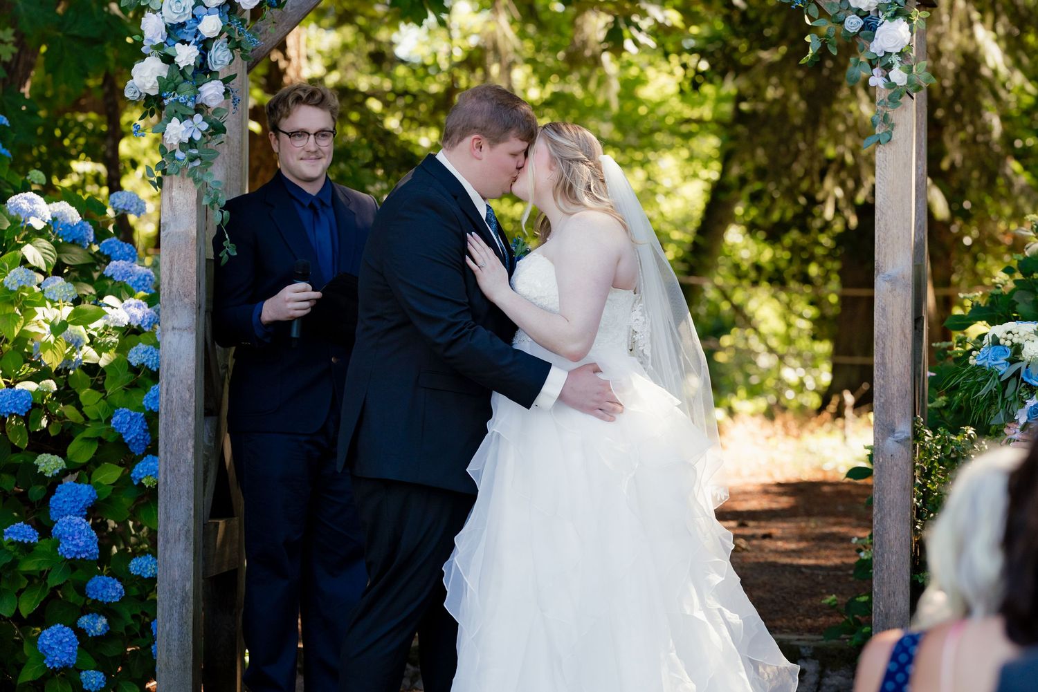 Bride and groom share their first kiss under a floral arch surrounded by blue hydrangeas at ceremony.