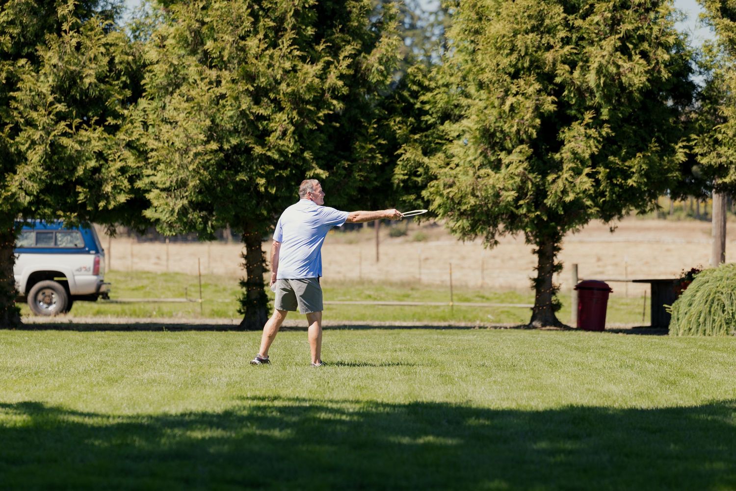 Man throwing a frisbee on a green lawn surrounded by tall trees on a sunny day.