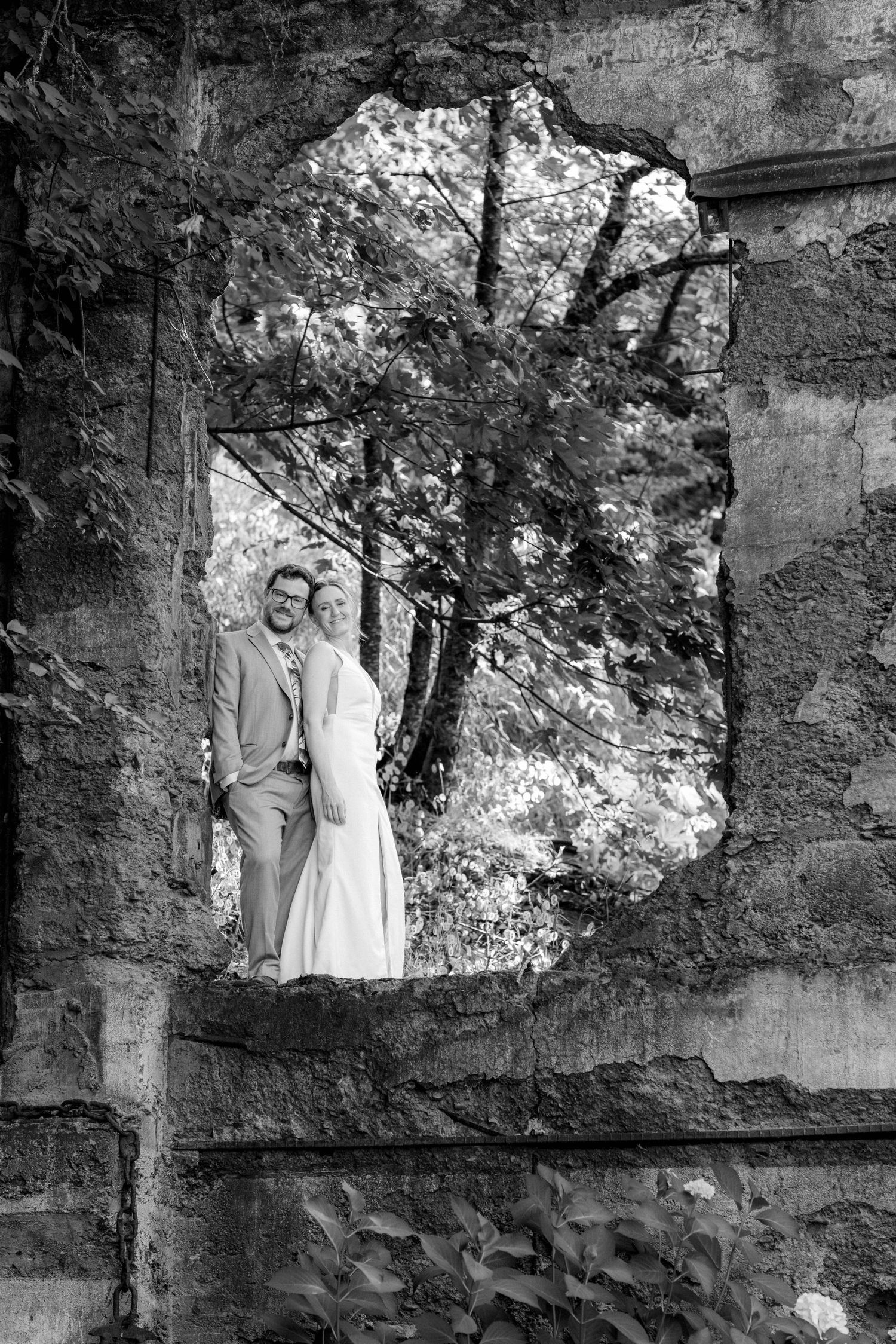 Black and white photo of bride and groom standing in a crumbling stone wall opening surrounded by lush trees.