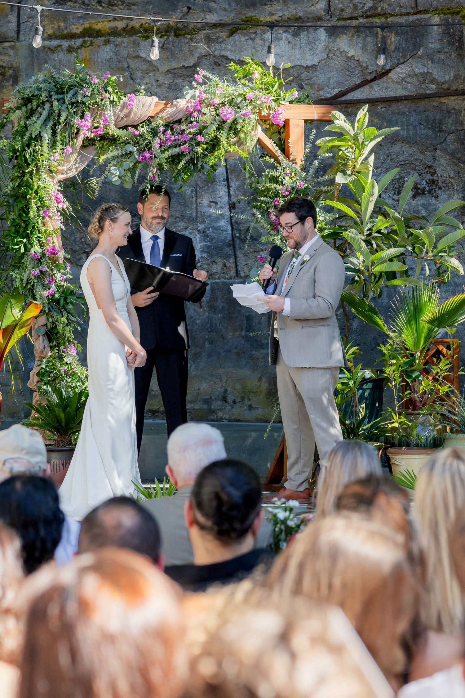 Bride and groom exchange vows under a floral arch with lush greenery at an outdoor wedding ceremony.