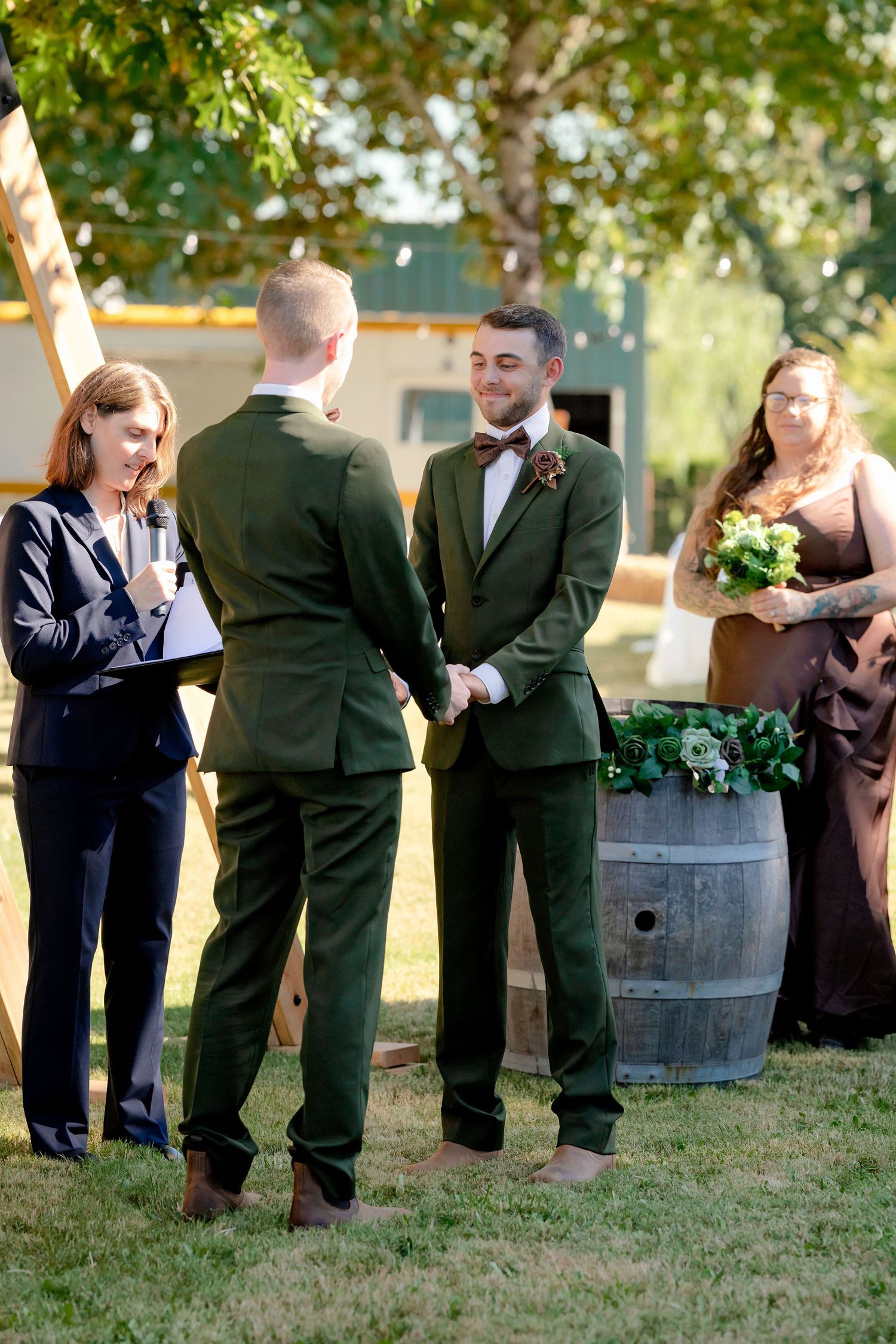 Two grooms in green suits holding hands during outdoor wedding ceremony with officiant.
