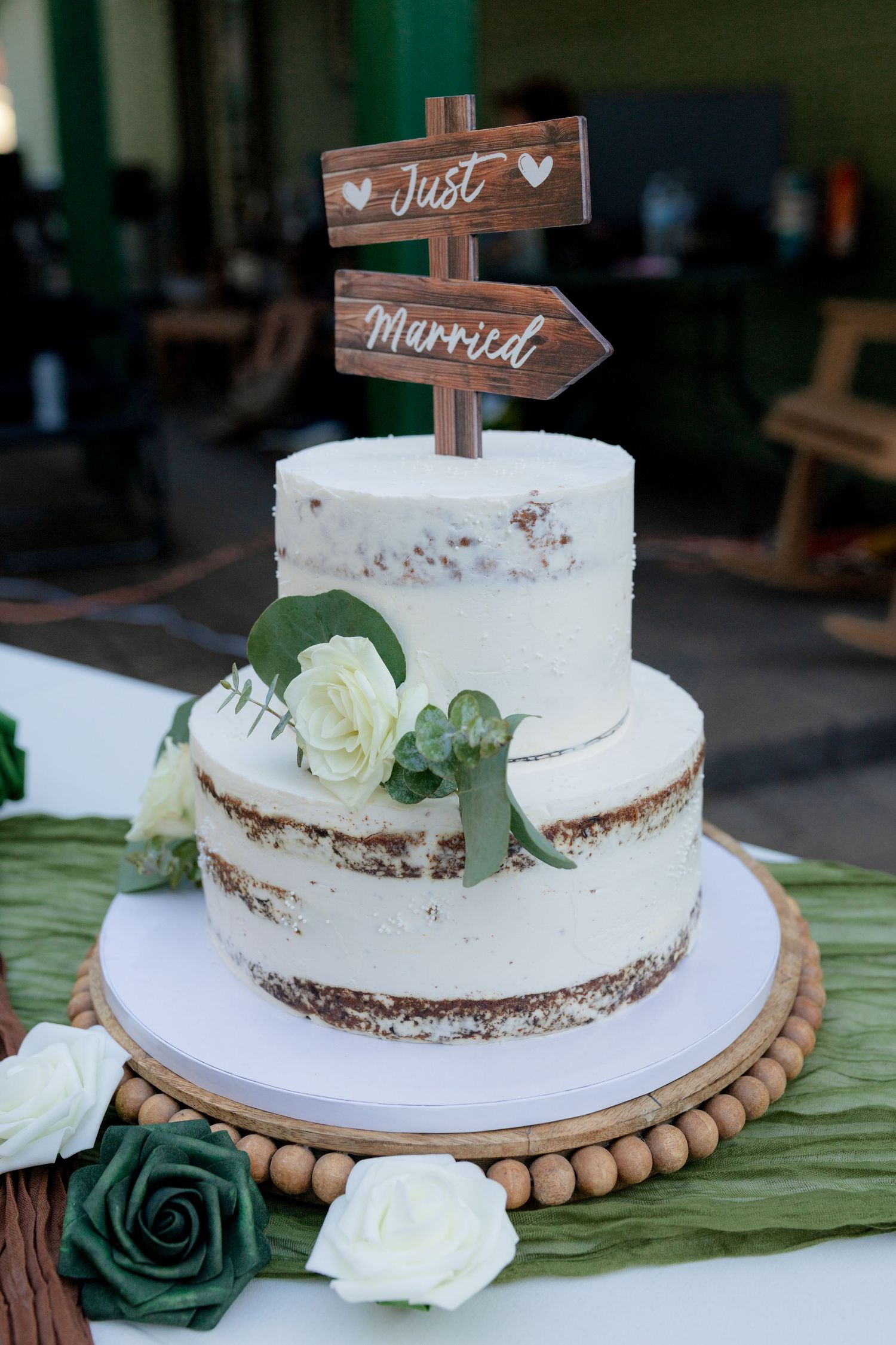 Two-tier naked wedding cake with white frosting and a wooden Just Married topper at a rustic wedding.
