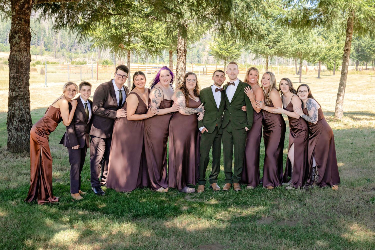 Wedding party posing together in brown and green attire under trees on sunny day.