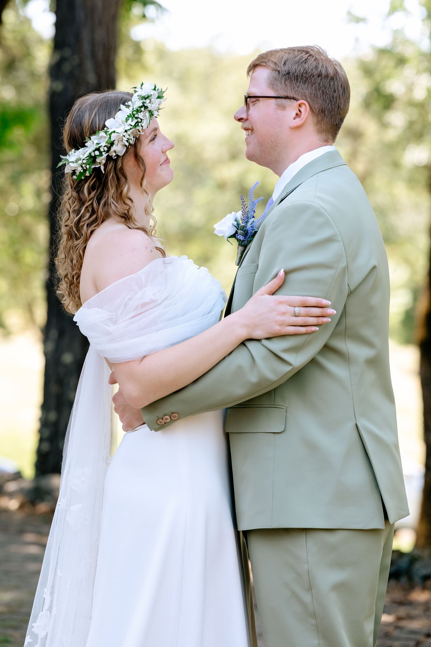 Bride in white gown and floral crown embracing groom in sage suit outdoors among trees.