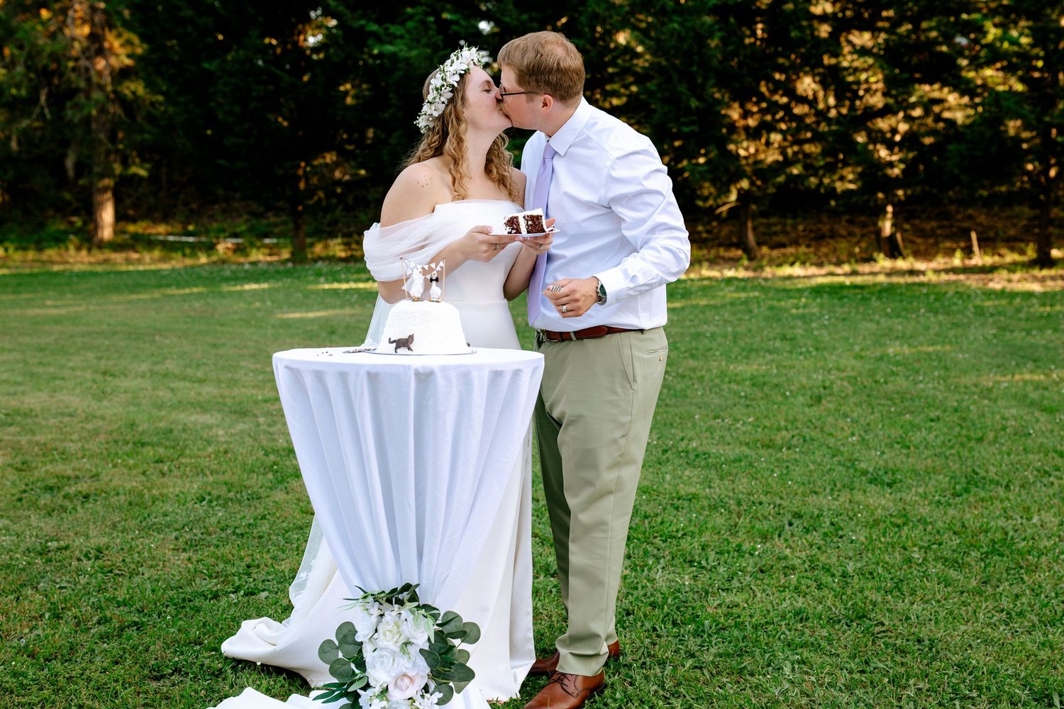 Newlyweds sharing a kiss while toasting champagne at a white cocktail table on a sunny green lawn.