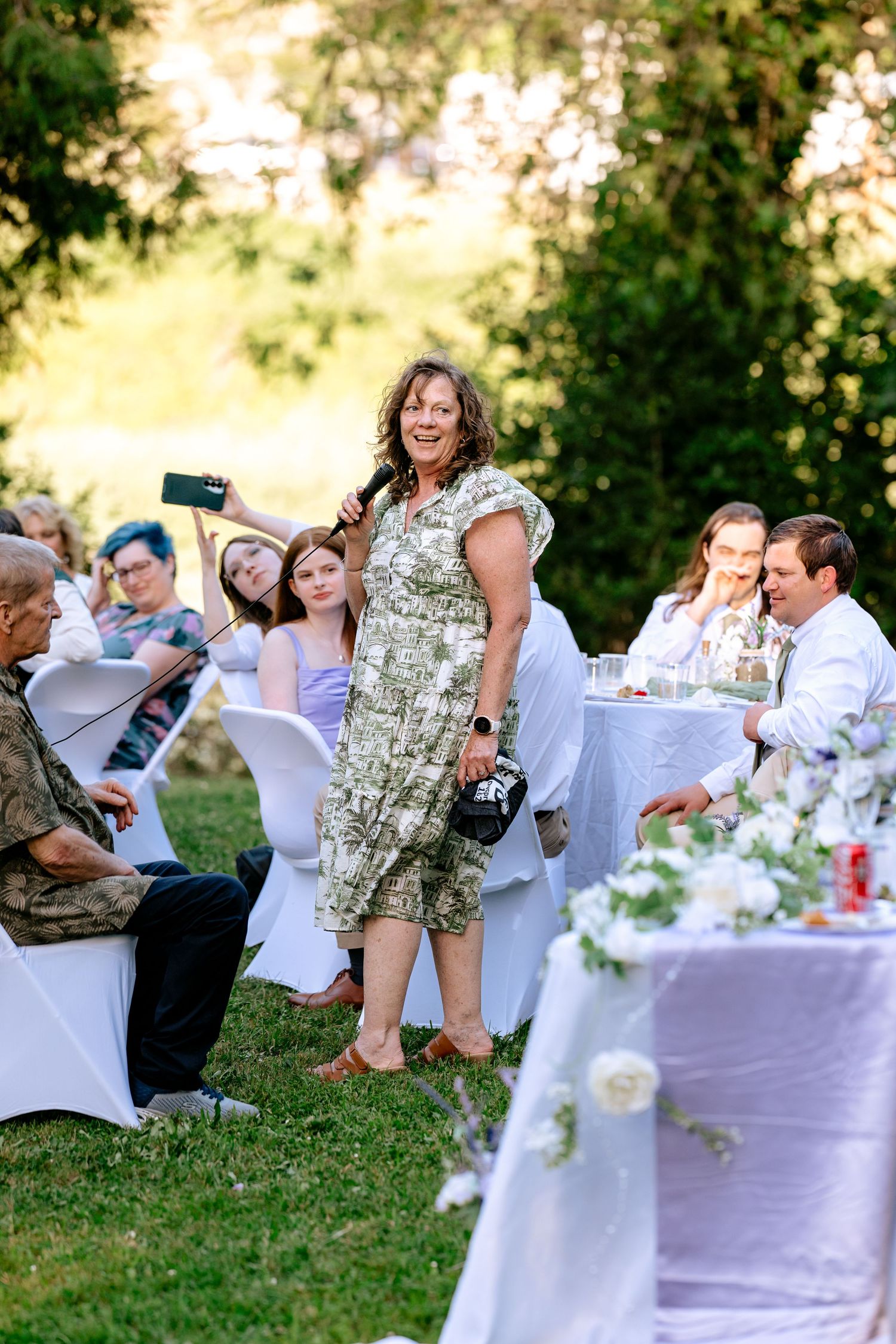 Woman in floral dress gives toast speech at elegant outdoor wedding reception on sunny evening.