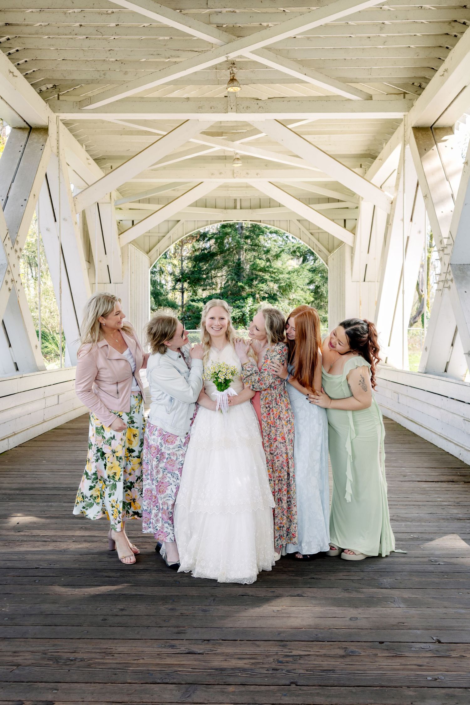 Bride in white gown holds bouquet surrounded by bridesmaids in floral dresses on a white covered wooden bridge.