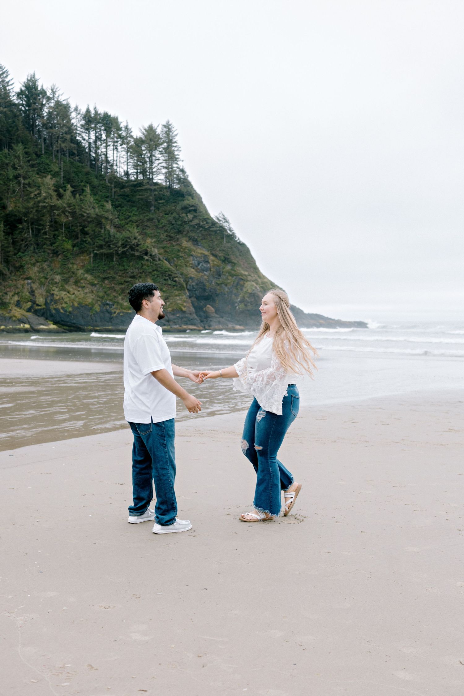Couple holding hands and dancing on sandy Pacific Northwest beach with forested cliffs.