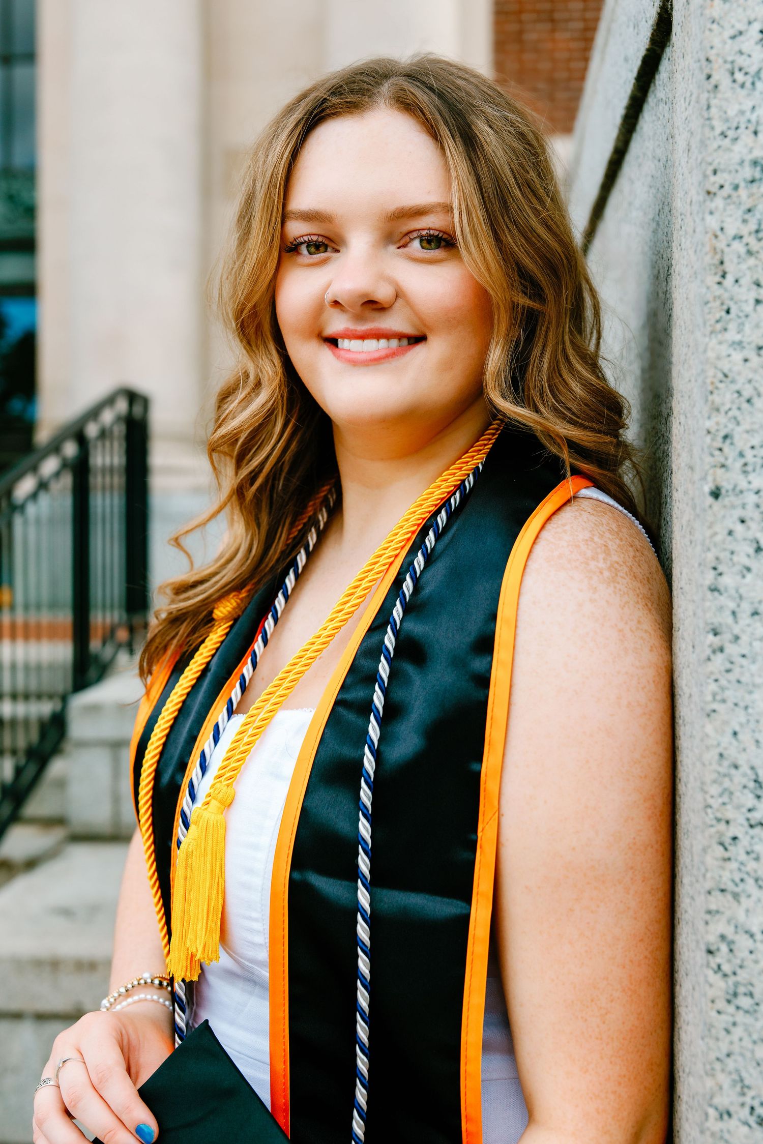 Smiling young woman in graduation stole with gold cords posing outdoors near stone column.