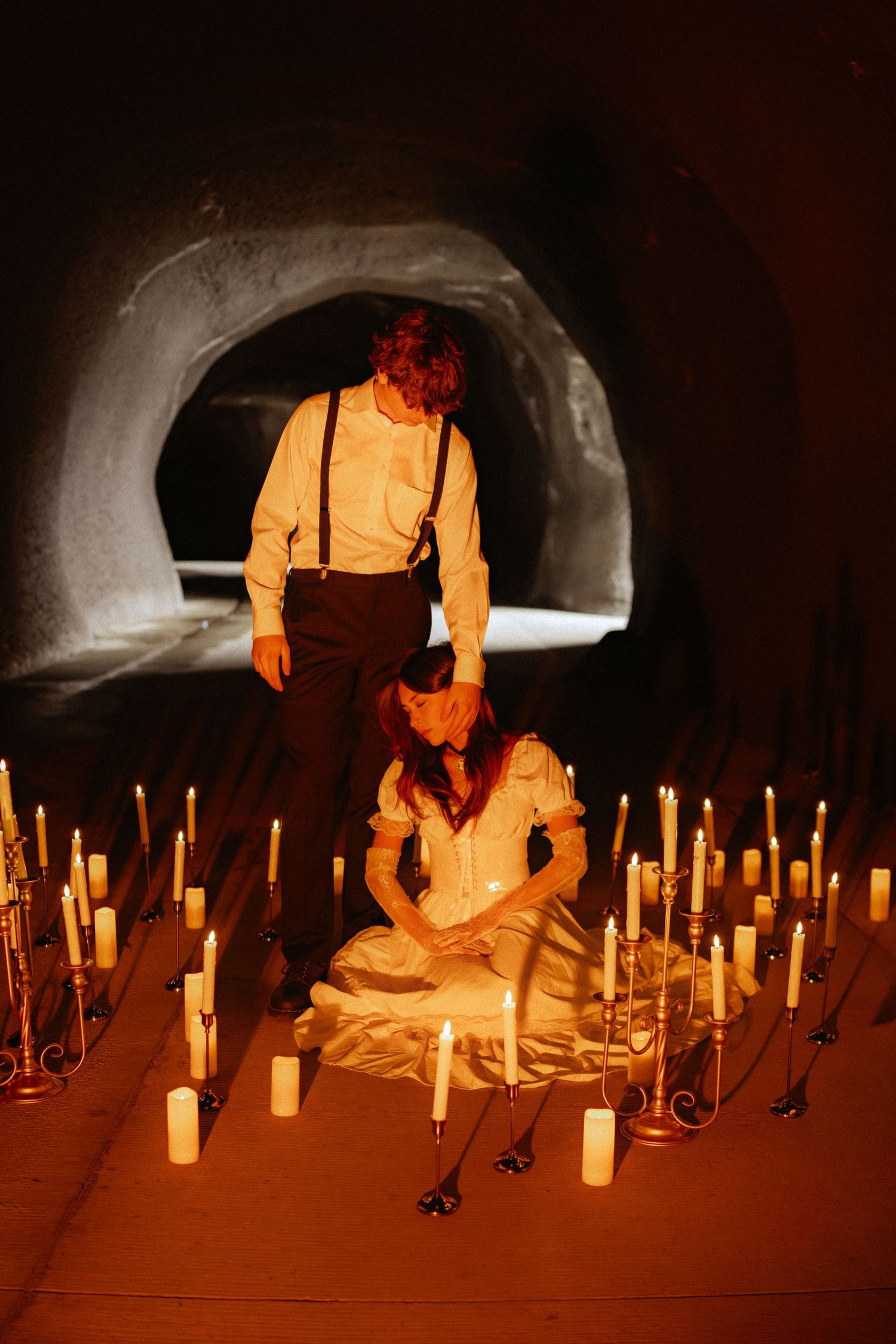 Two figures in vintage attire surrounded by lit candles in a dark tunnel setting create an eerie gothic scene.