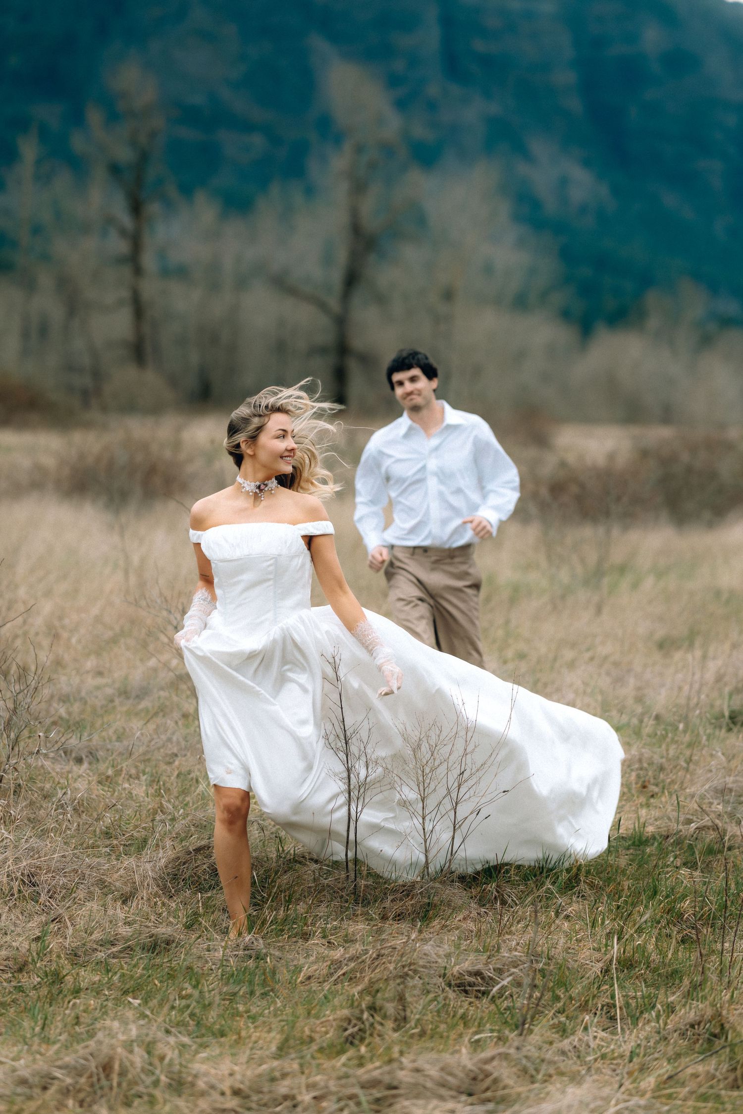 A couple in wedding attire playfully falls together in a rustic field with tall grass and moody lighting.